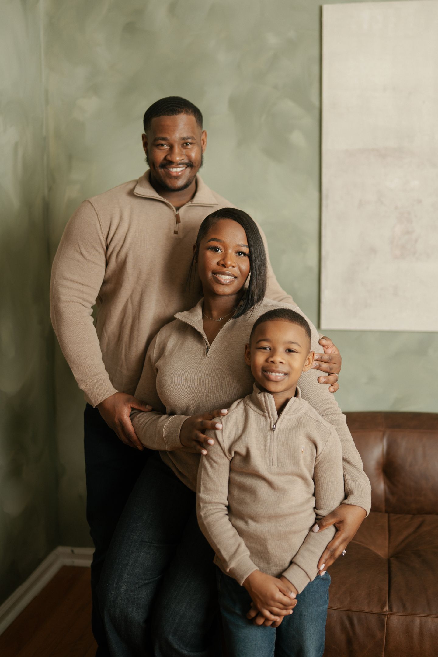A family wearing matching beige sweaters poses together against a neutral backdrop with a leather couch.