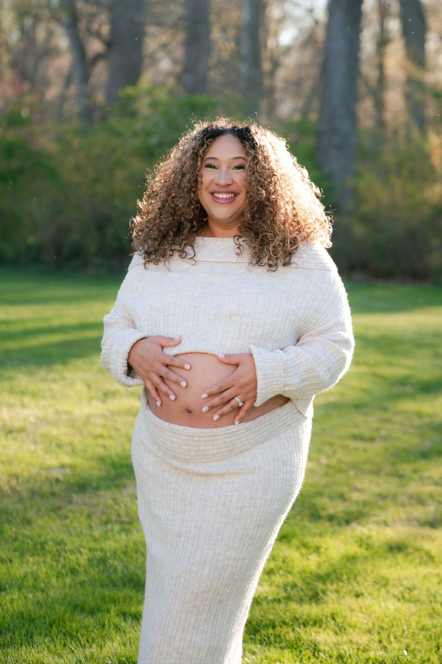 Person in white sweater poses joyfully on grass during outdoor photoshoot.