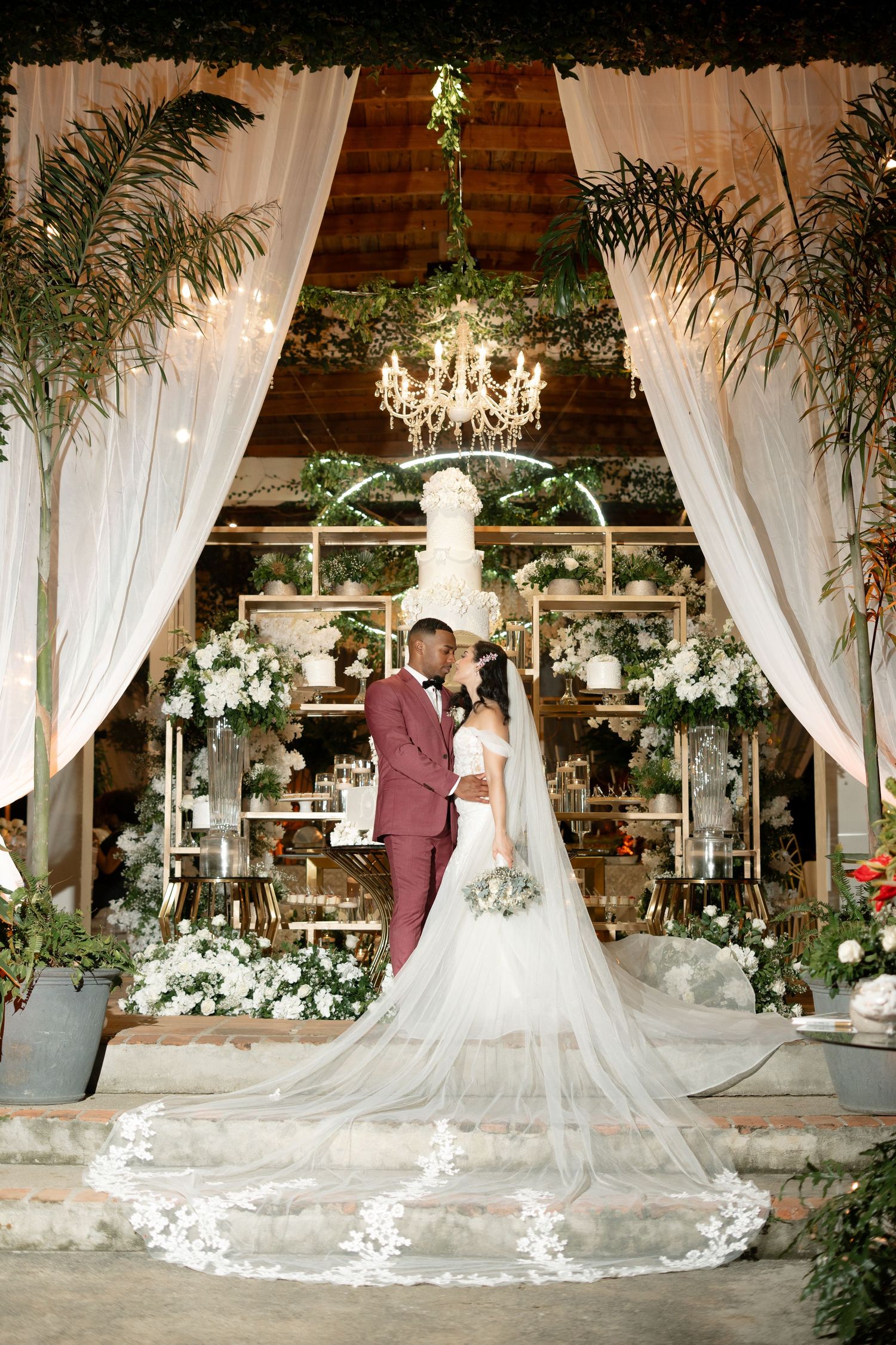 Elegant indoor wedding venue decorated with white drapes, chandeliers and palm trees.