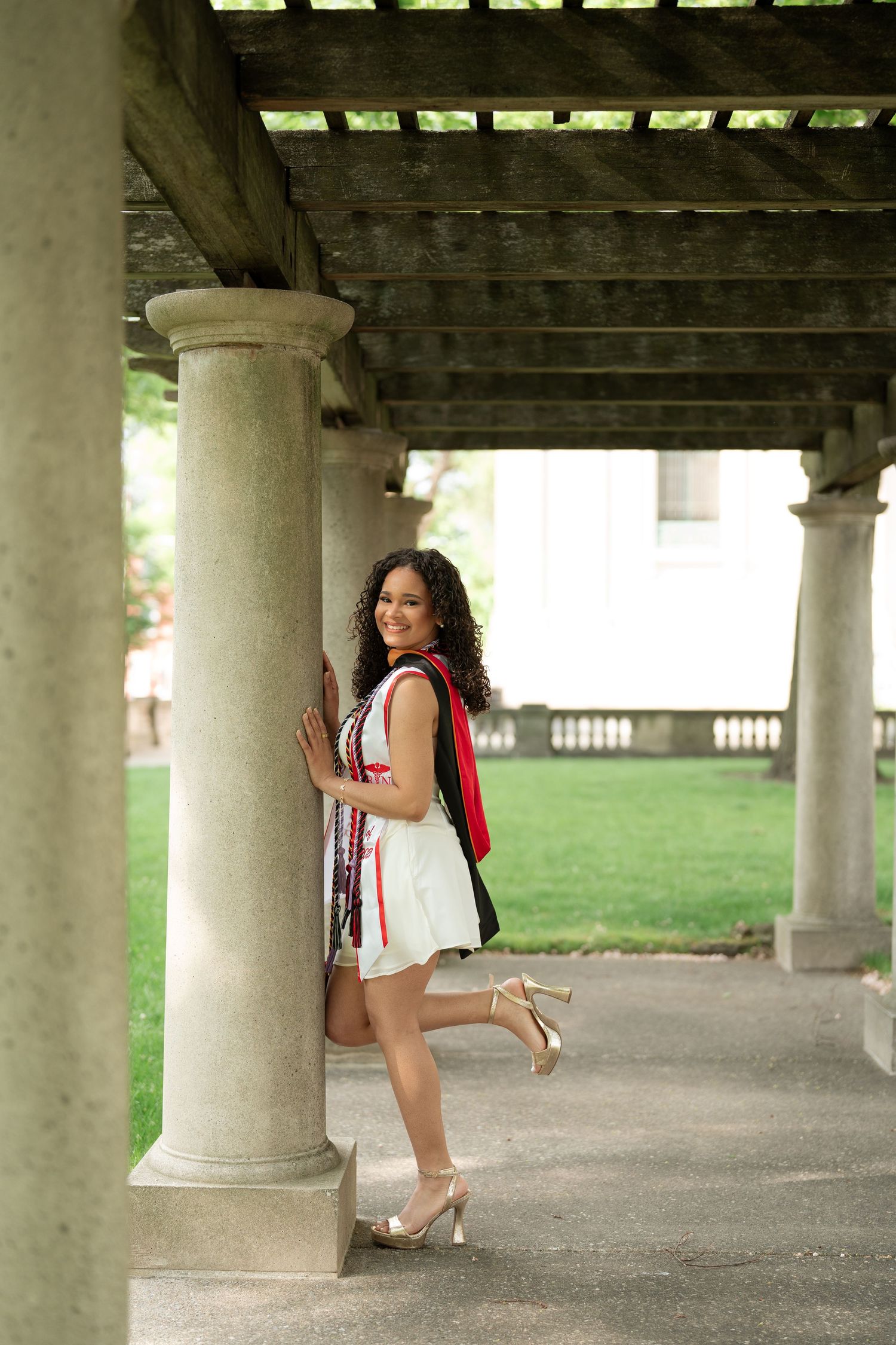 Young woman in white dress and heels leans against stone column under pergola during graduation photoshoot.