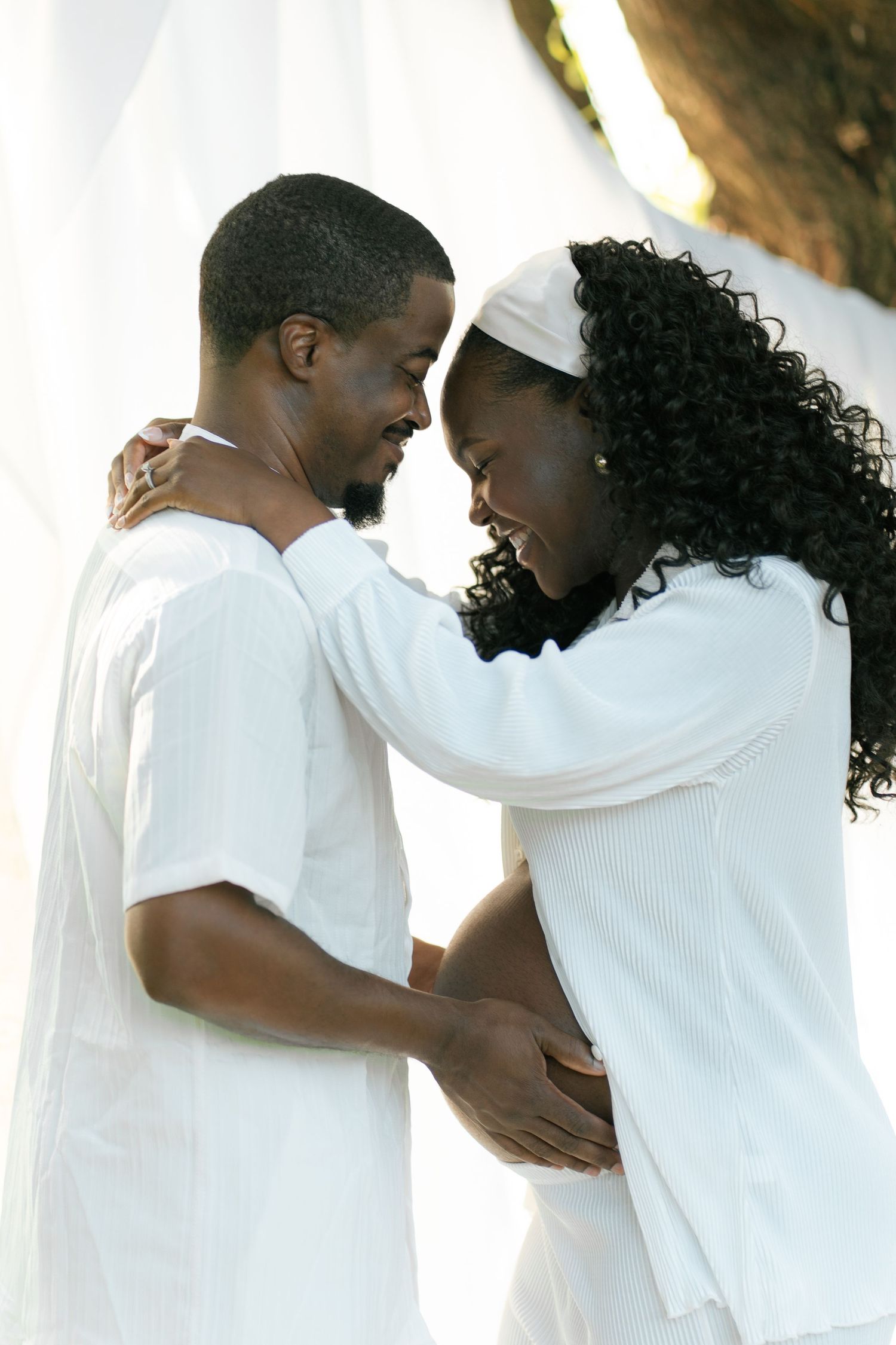 A loving Black couple in matching white outfits embrace tenderly, foreheads touching, outdoors near a tree.