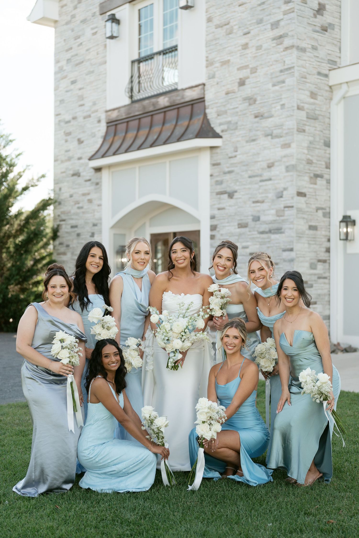Bride with bridesmaids in mismatched blue satin dresses holding white bouquets outside stone wedding venue.