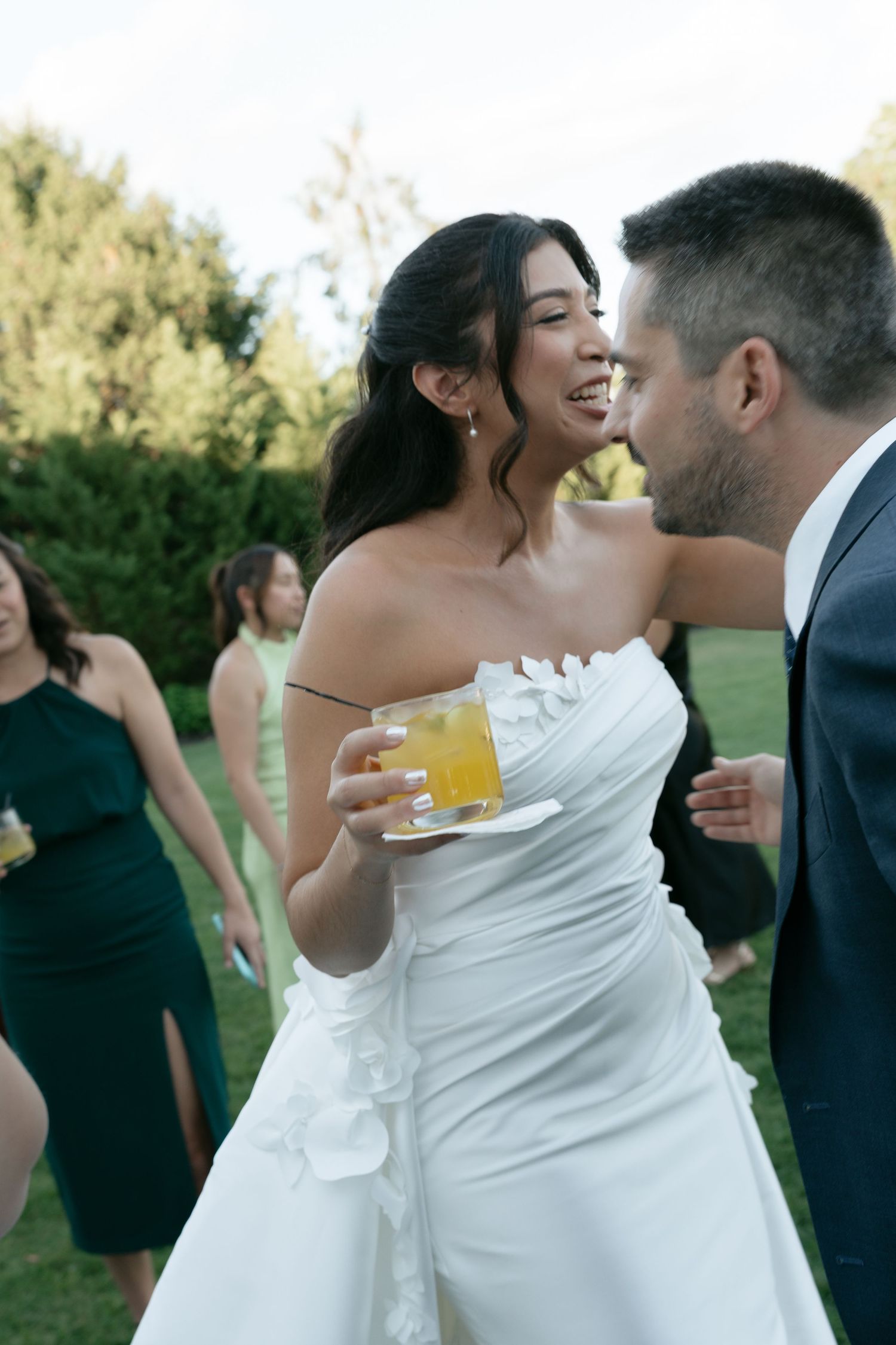 Smiling bride in white gown holds cocktail while chatting with groom outdoors.