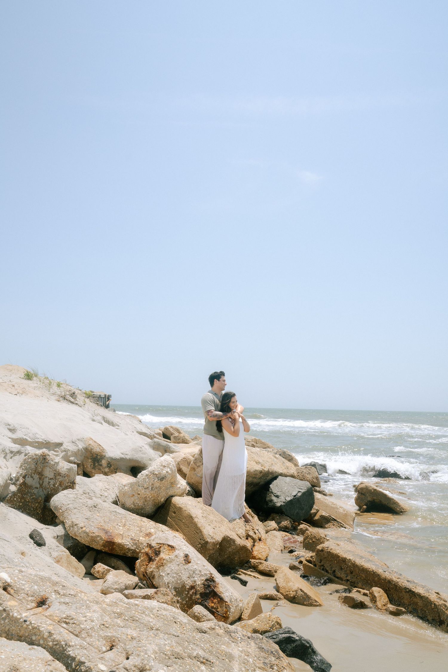 A person sits on coastal rocks overlooking the ocean waves crashing against the shore on a sunny day.