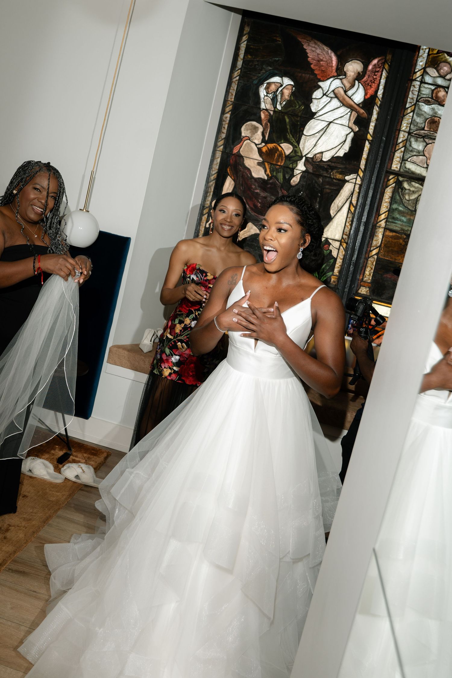 Joyful wedding preparation moment with bride and friends against stained glass backdrop.