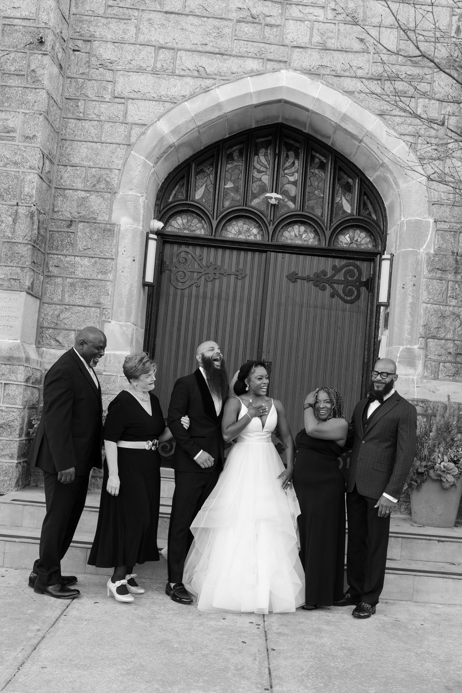 Wedding party shares candid moment outside church entrance in black and white photography.