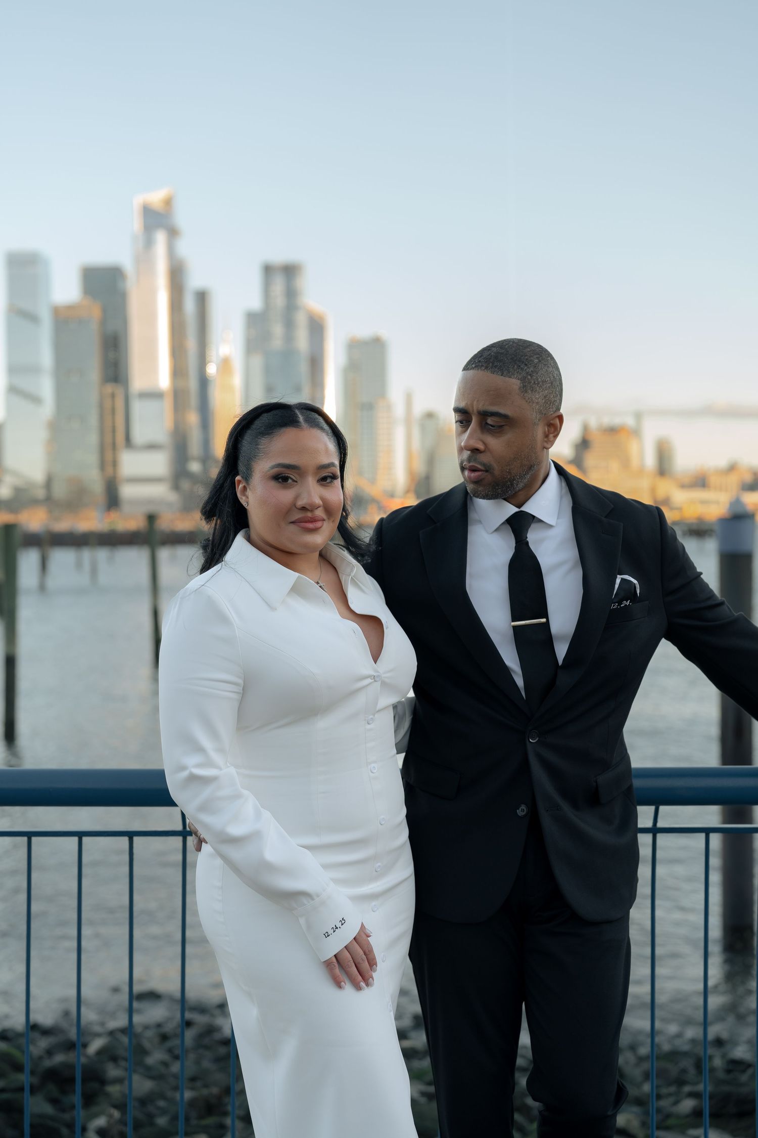A couple poses together on a waterfront with a dramatic city skyline in the background.