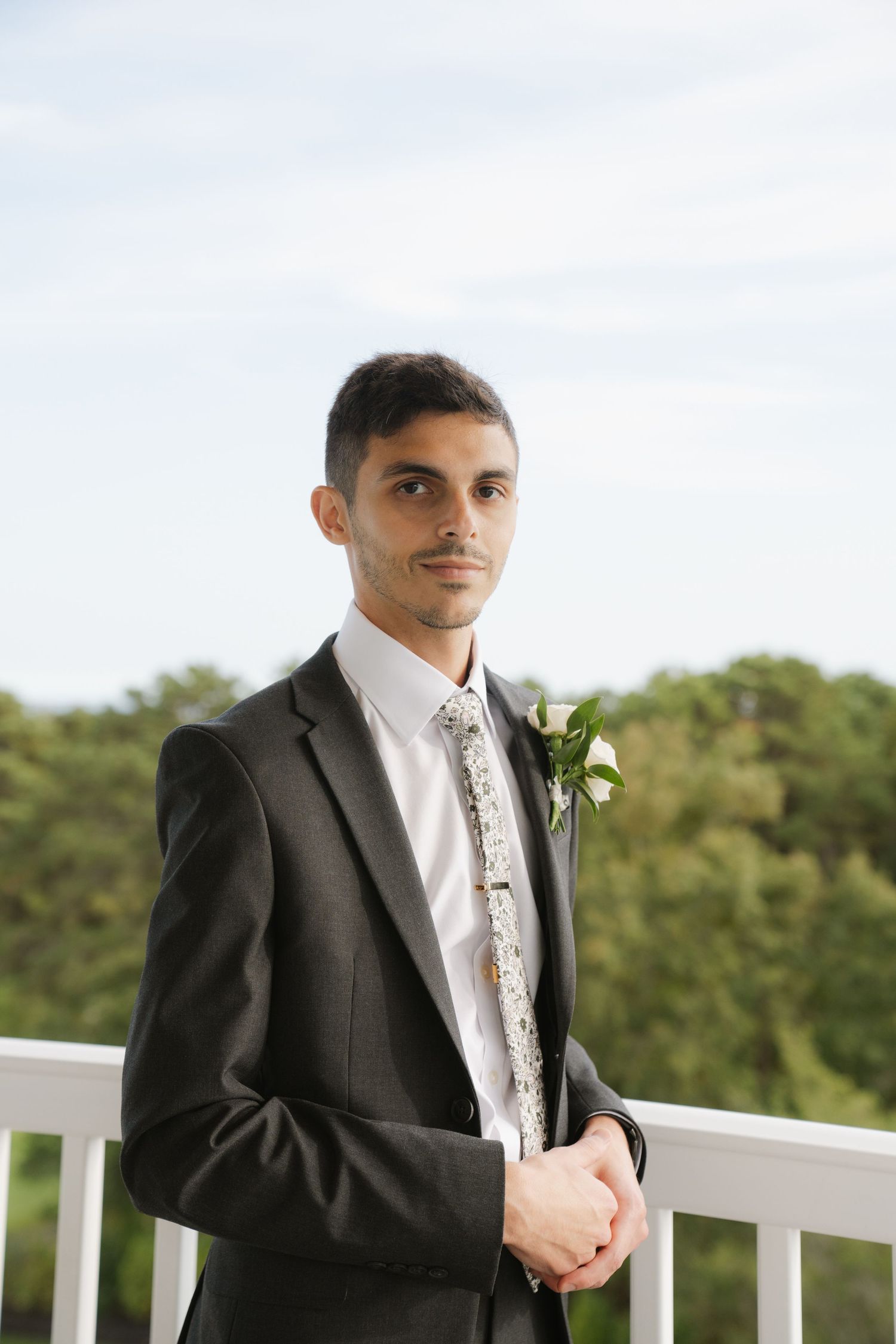 A groom in a black suit and floral tie stands on a white balcony railing against a green outdoor background.