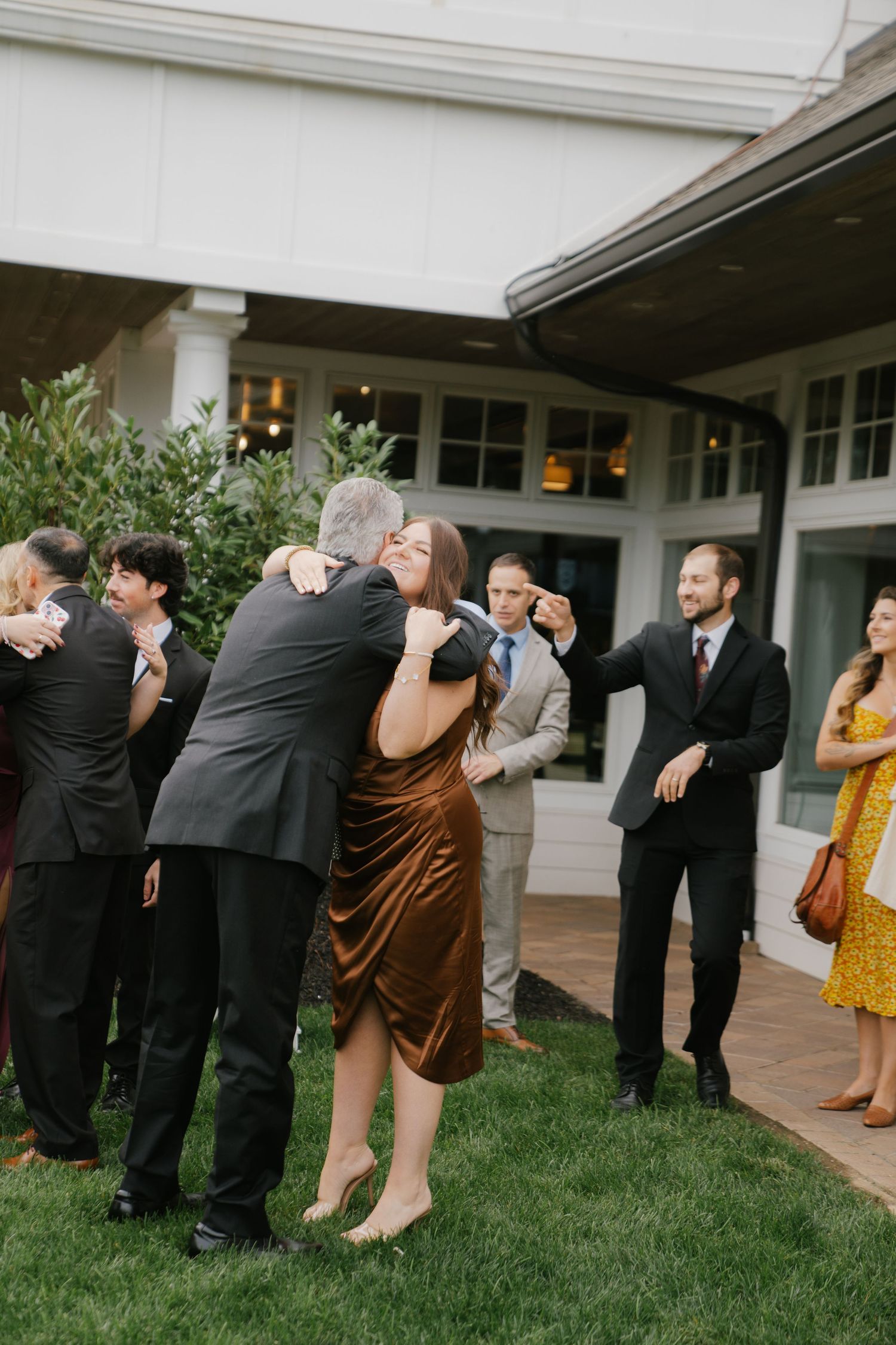 Wedding party members in black suits and dresses perform a synchronized dip dance move on grass outside a venue.