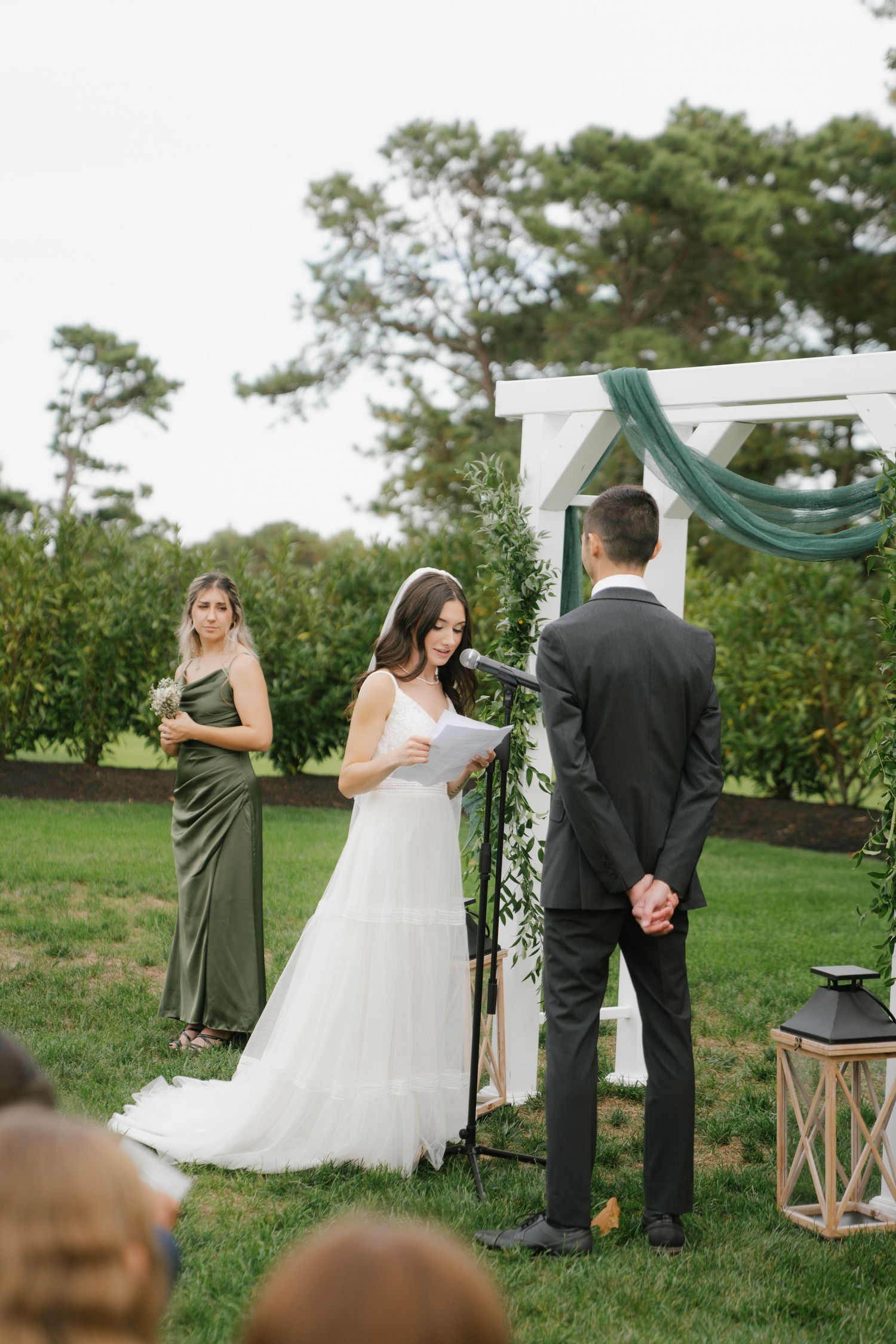 An outdoor wedding ceremony takes place under a white arch with green drapery and floral decorations on a grassy lawn.