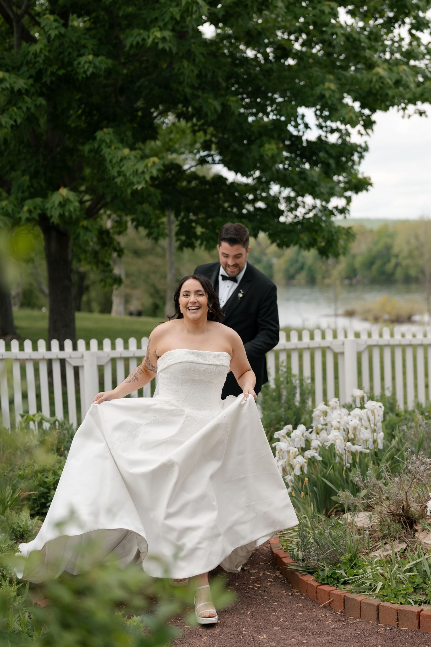 A joyful bride in a strapless white gown laughs as her groom in a black tuxedo helps carry her dress train in a garden.