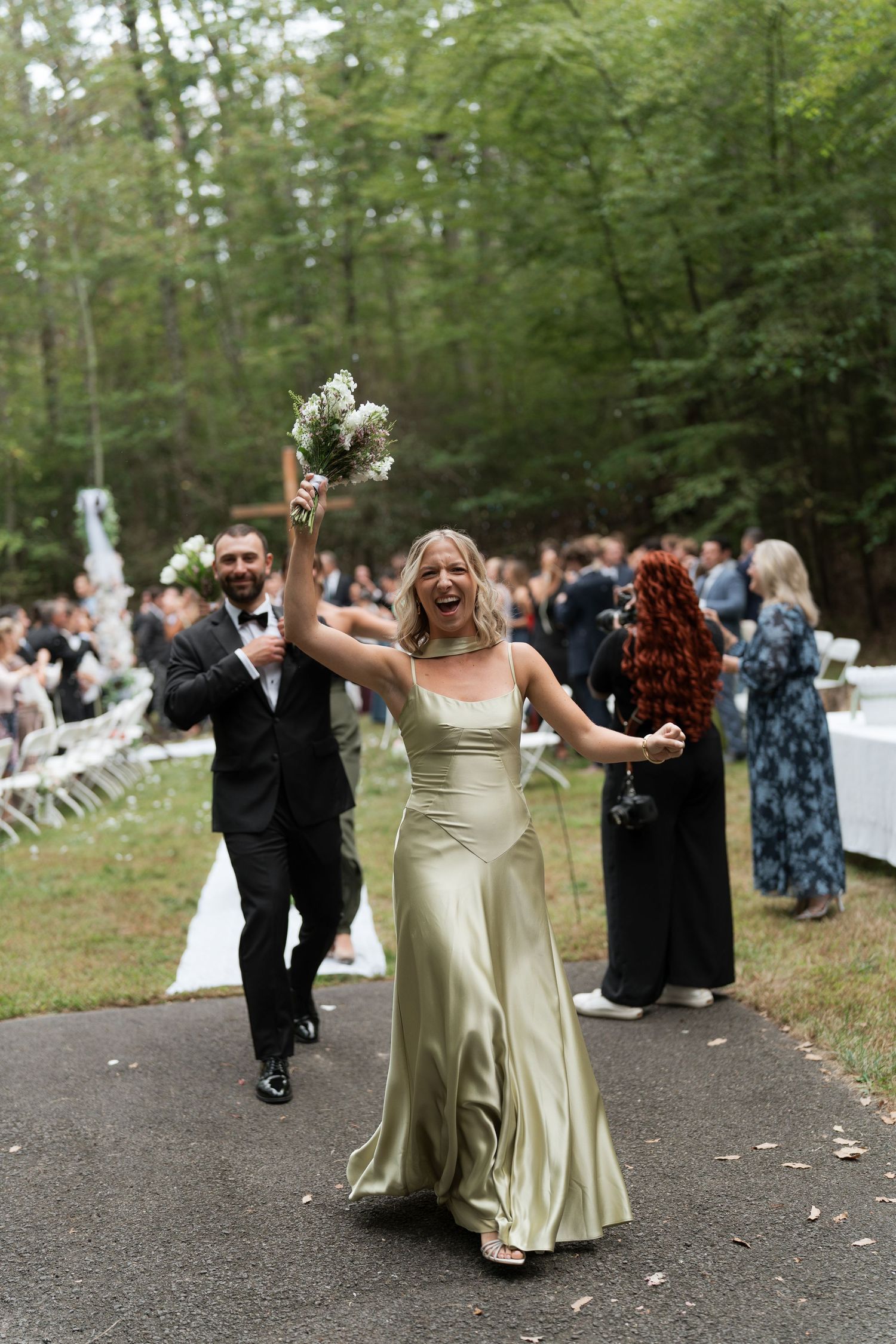 Joyful bridesmaid in sage green satin gown raises bouquet celebrating after outdoor wedding ceremony.