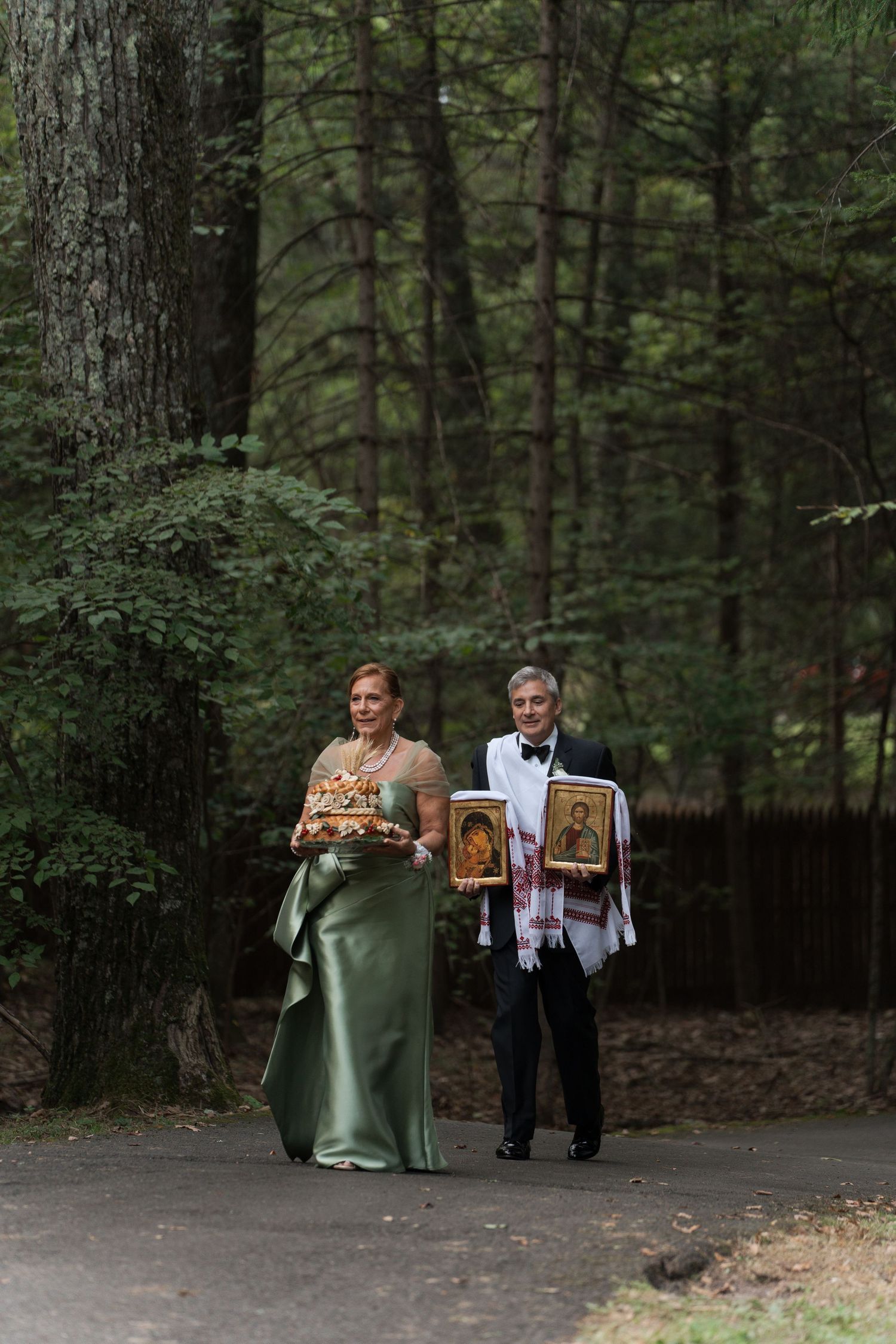 Woman in green gown and man in tuxedo carry Orthodox wedding icons through a forest path.