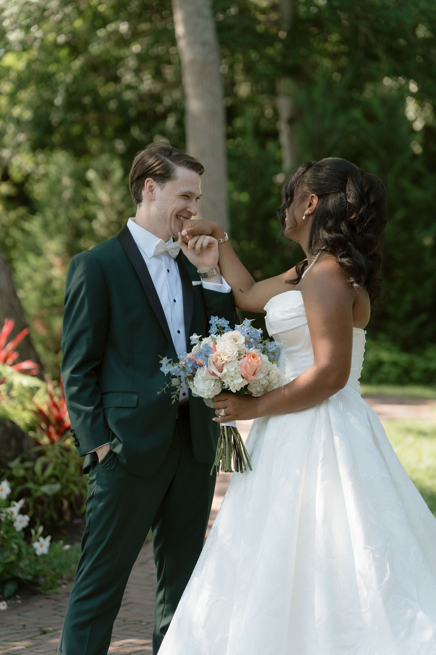 Interracial couple shares a tender moment outdoors, bride in strapless gown holding pastel bouquet, groom in dark green tuxedo.