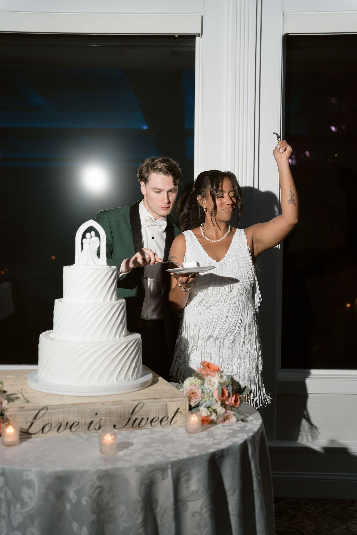 Bride and groom cut four-tier white wedding cake at table decorated with Love is Sweet sign.