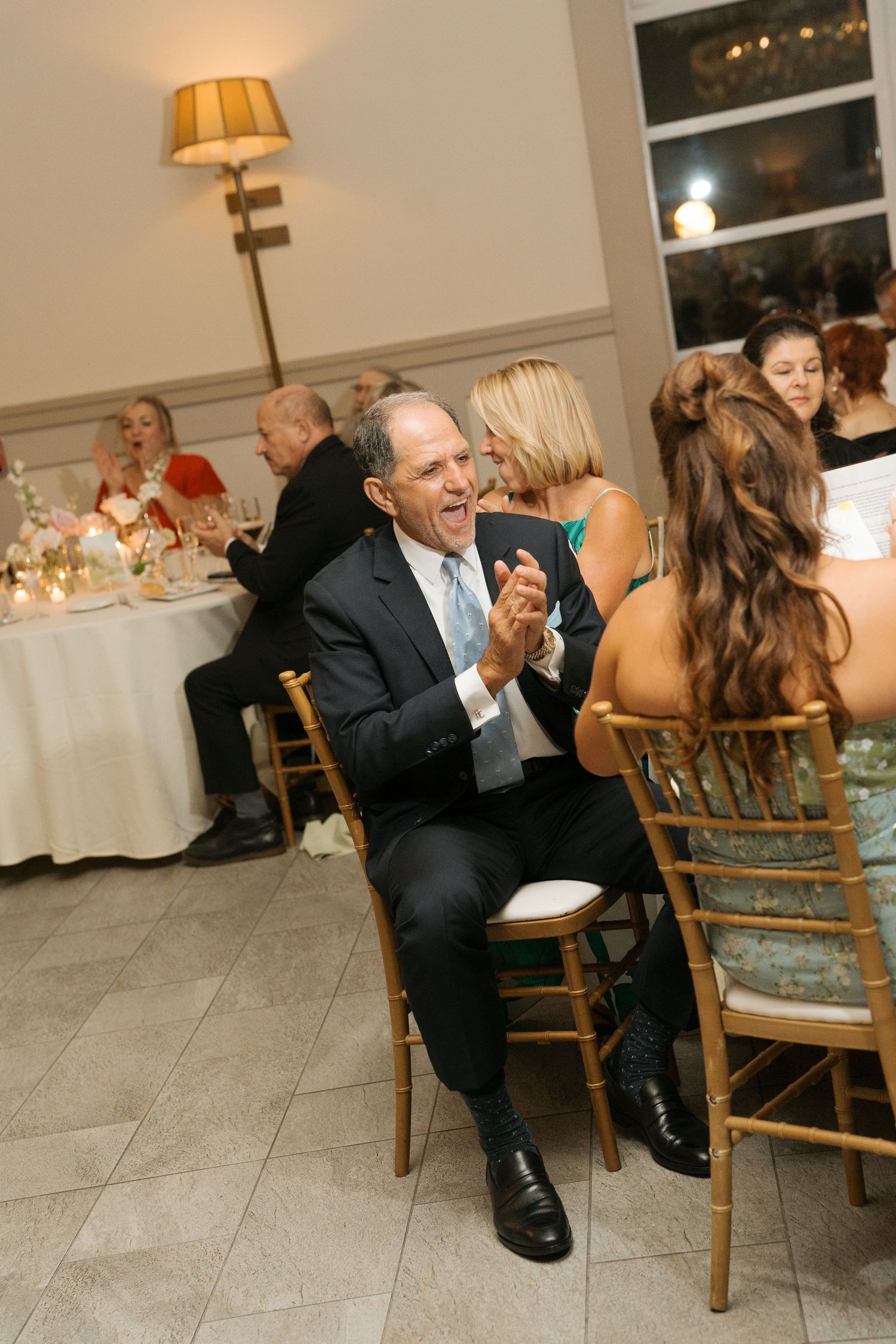 Elderly man in a suit laughing and clapping while seated at a wedding reception dinner table.
