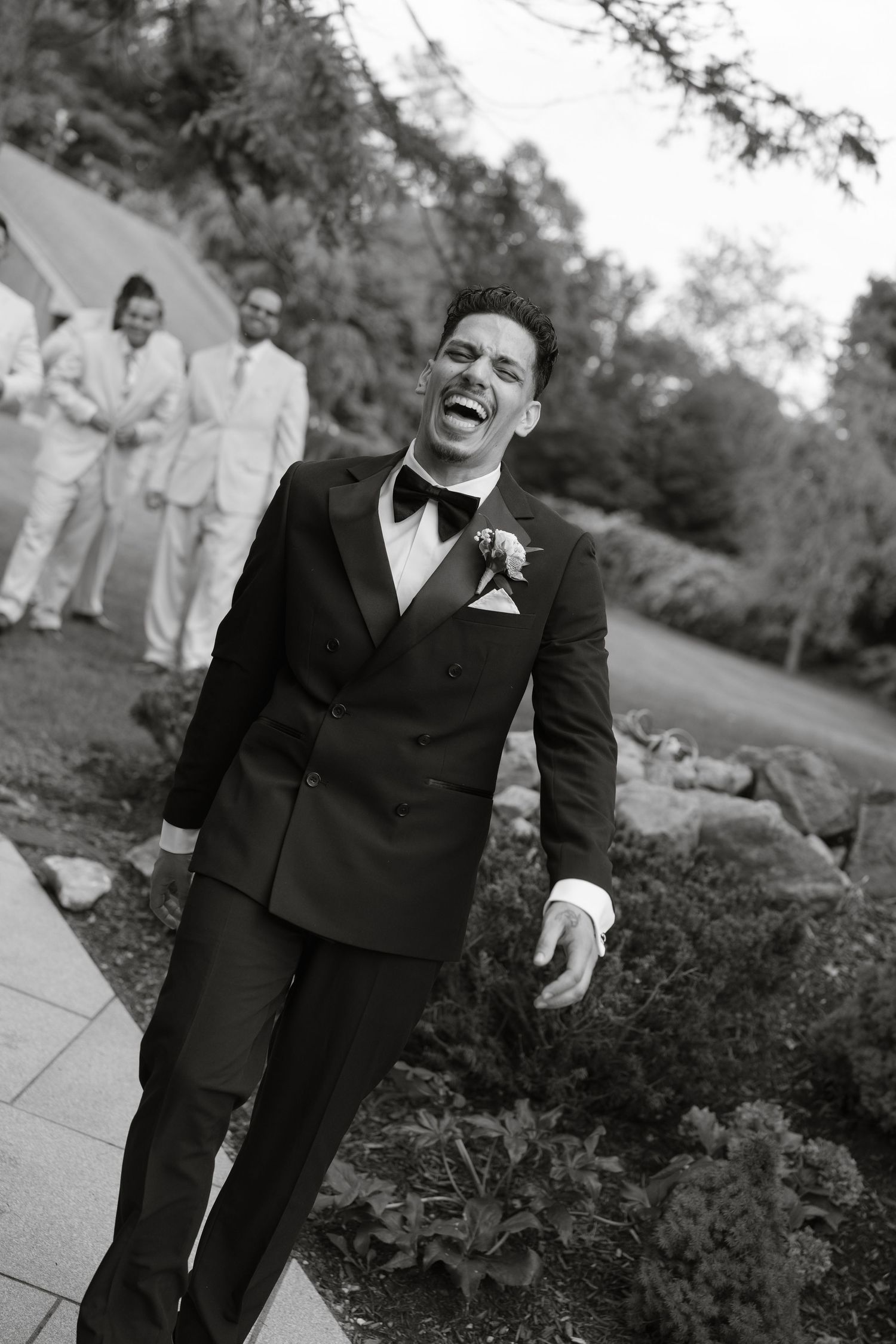Groom laughing joyfully in black tuxedo with boutonniere, groomsmen in white suits visible in background.