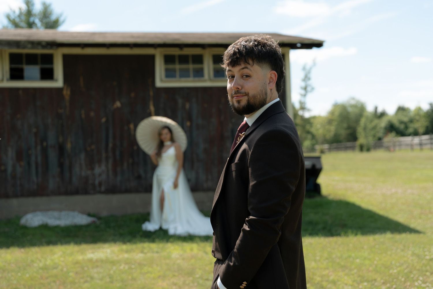 Groom in black suit stands in foreground while bride in white gown and sun hat poses near rustic barn outdoors.