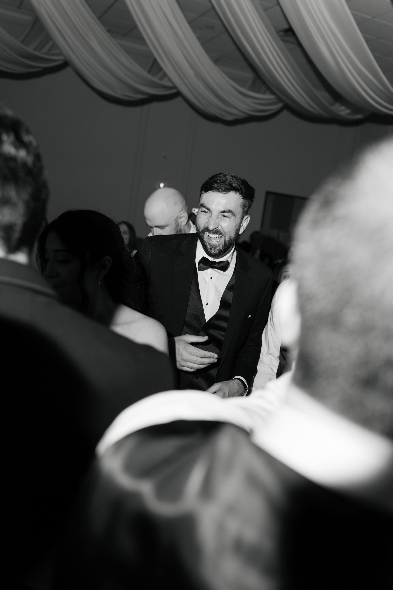 Black and white photo of smiling groom in tuxedo dancing with guests at wedding reception.