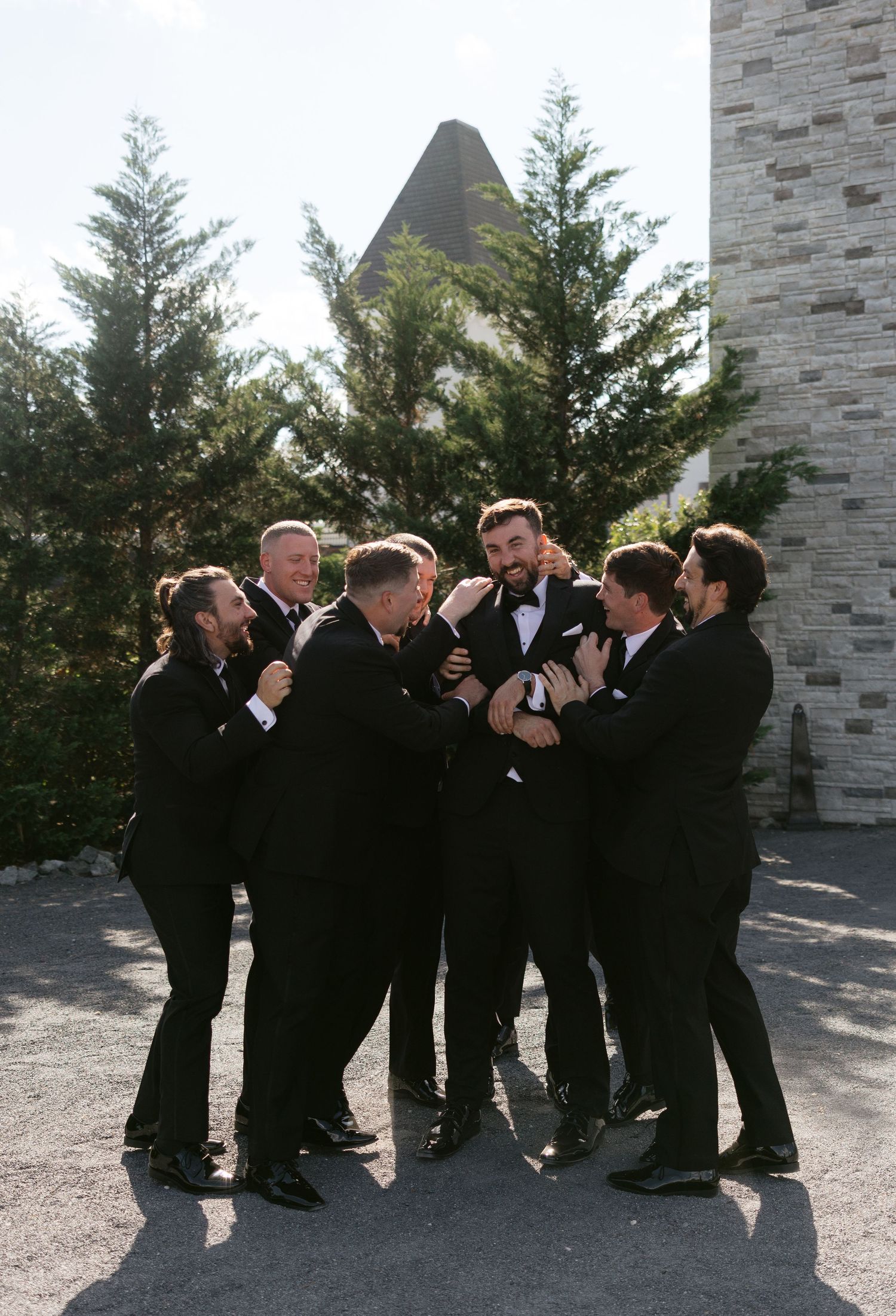 Groom surrounded by groomsmen all in black tuxedos playfully celebrating outside stone wedding venue.