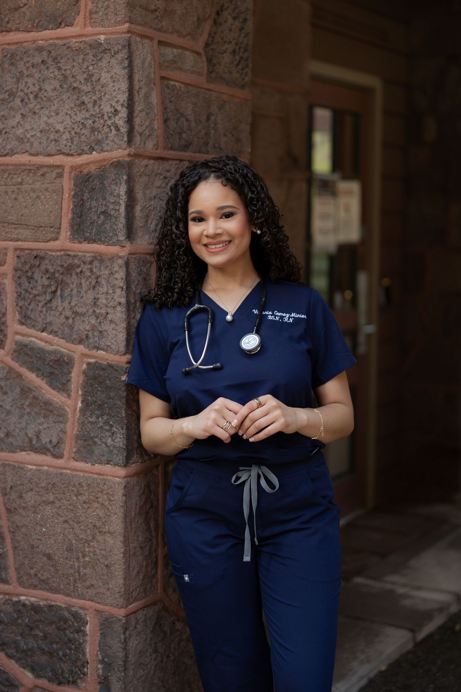 Female nurse in navy scrubs with stethoscope leaning against stone wall smiling outdoors.