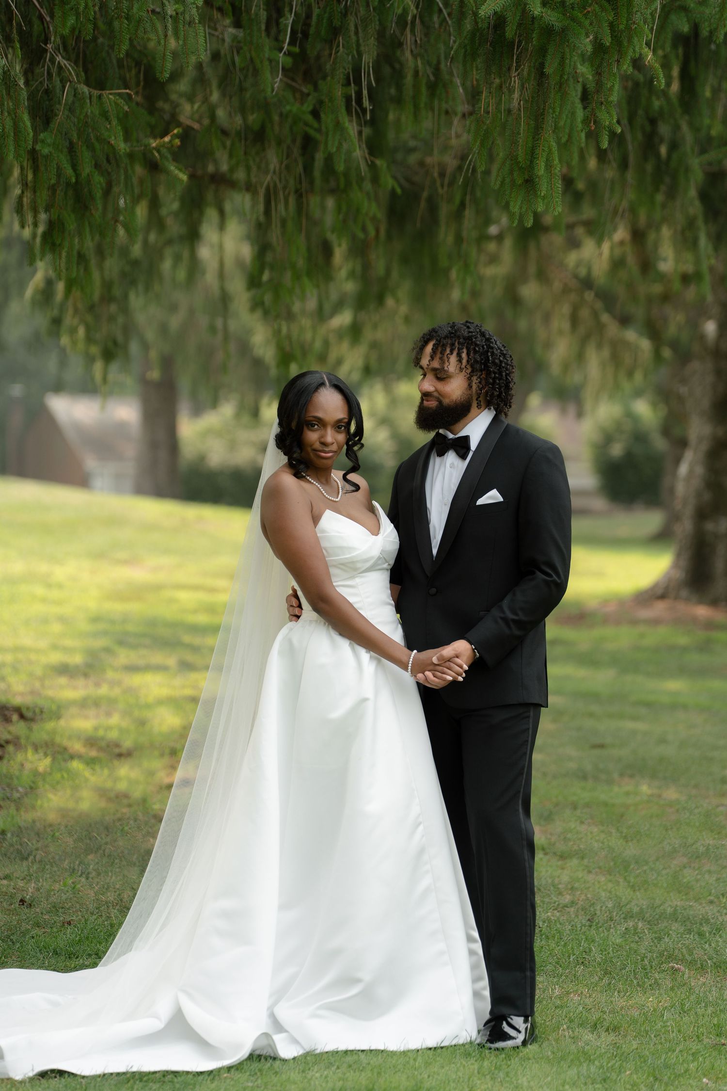 Elegant bride in white gown and groom in black tuxedo posing together under a weeping willow tree.