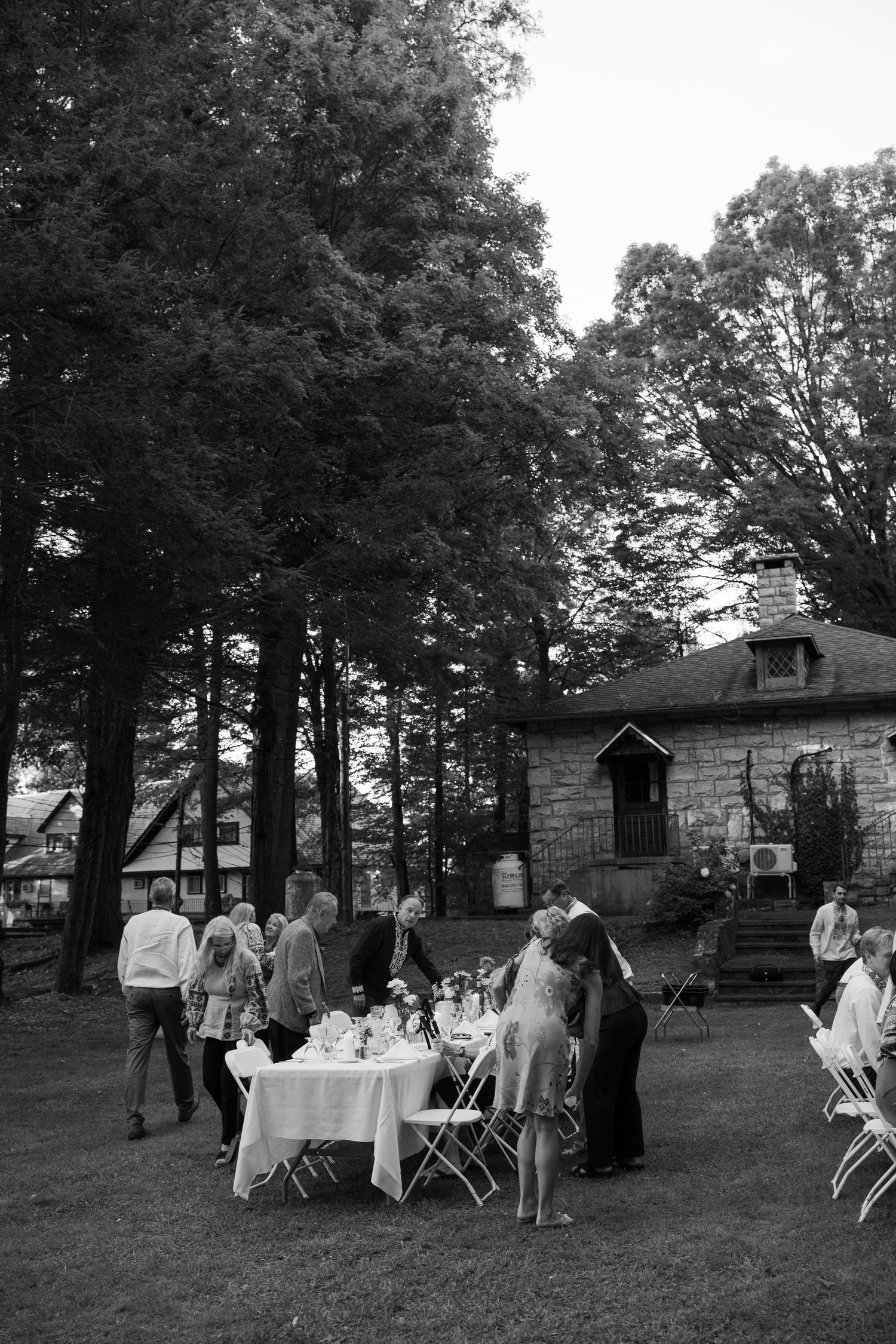 Black and white photo of wedding guests gathered around an outdoor reception table beneath tall trees near a stone building.