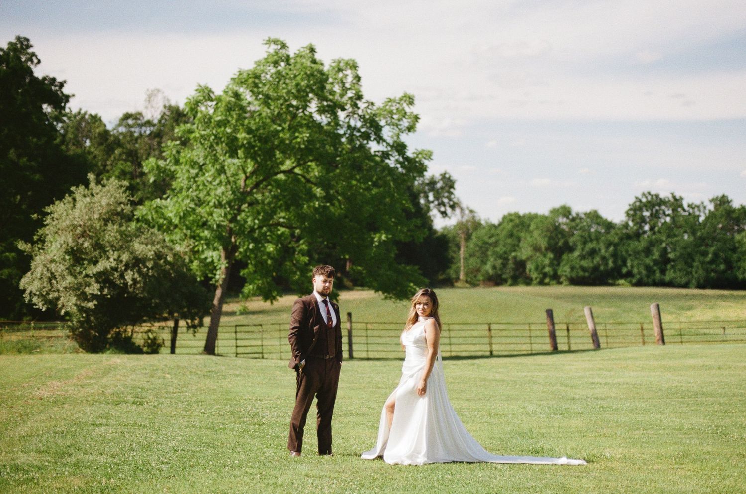 Bride and groom stand apart in a lush green field with wooden fence and trees at an outdoor wedding.