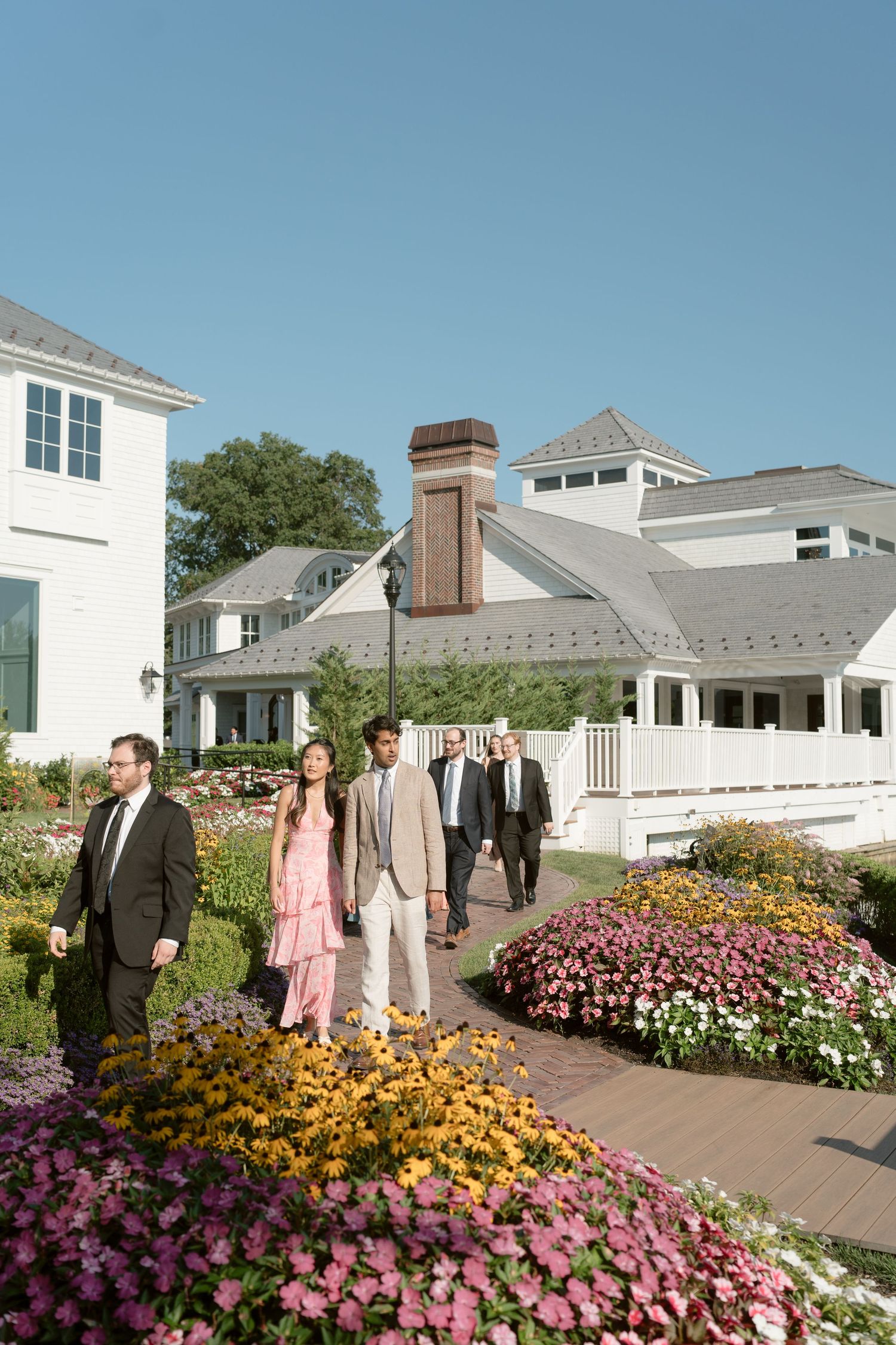 Wedding guests in formal attire walk through a vibrant flower garden toward a white venue on a sunny day.