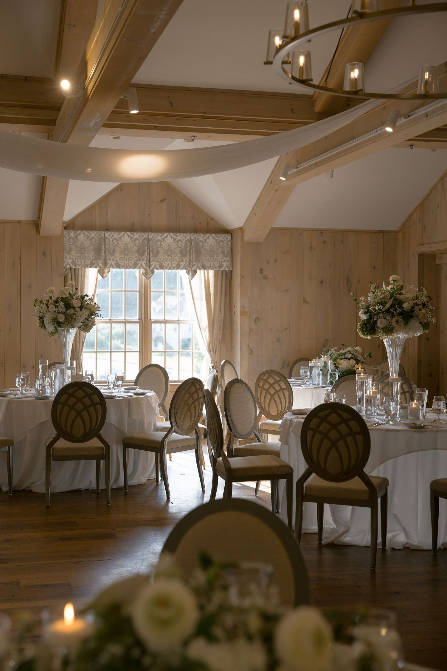 Rustic wedding reception venue with vaulted wood ceiling, round tables, elegant chairs, floral centerpieces, and candlelight.
