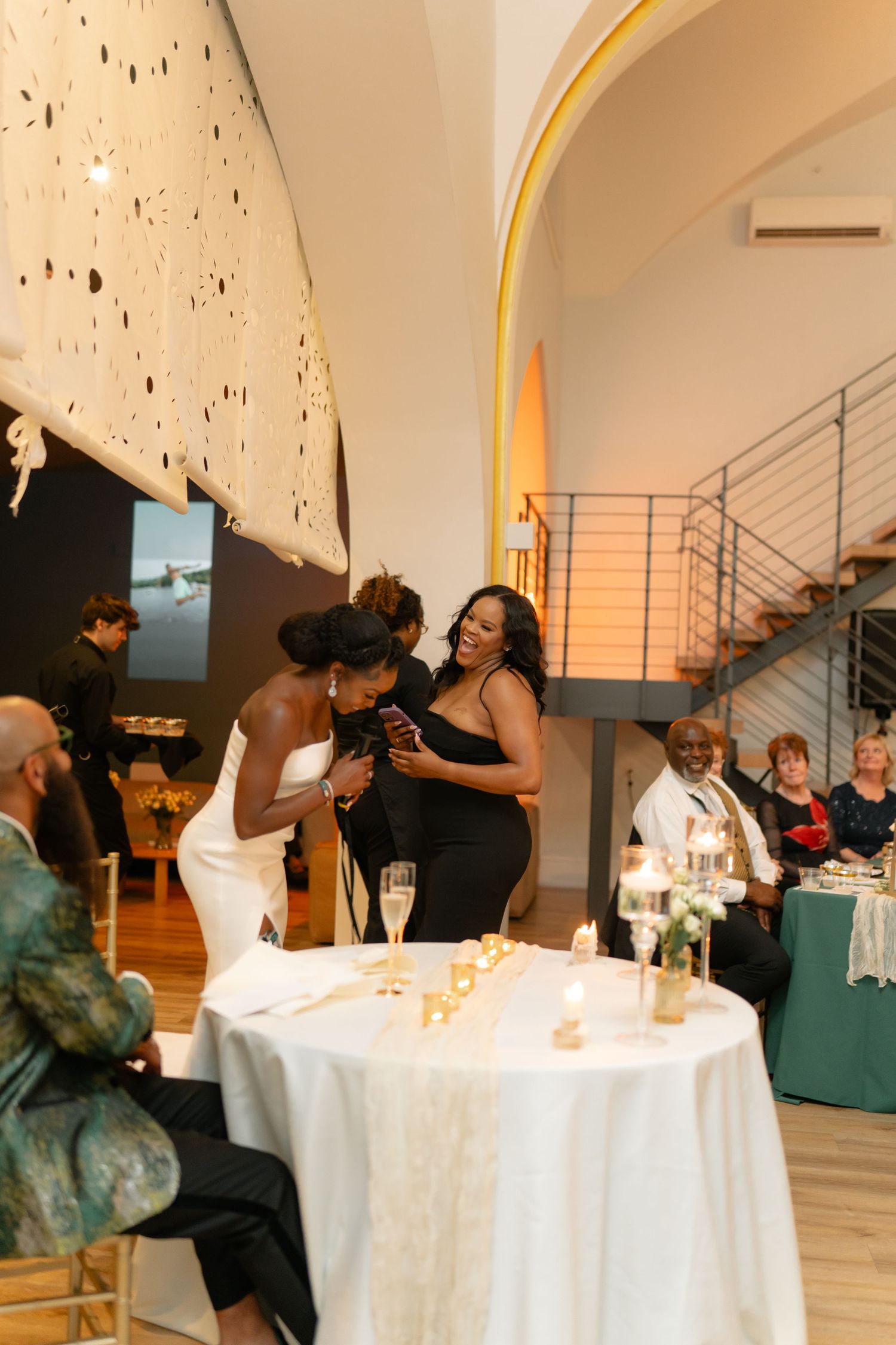 Wedding guests celebrate at round tables with white tablecloths under an arched ceiling.