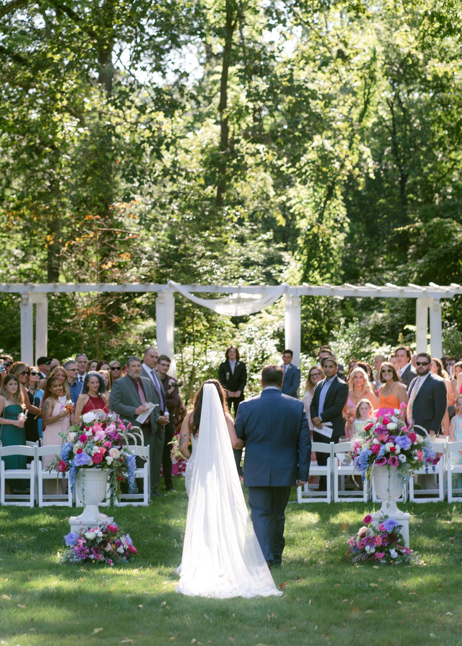 Outdoor wedding ceremony setup with white chairs and floral arrangements under trees.
