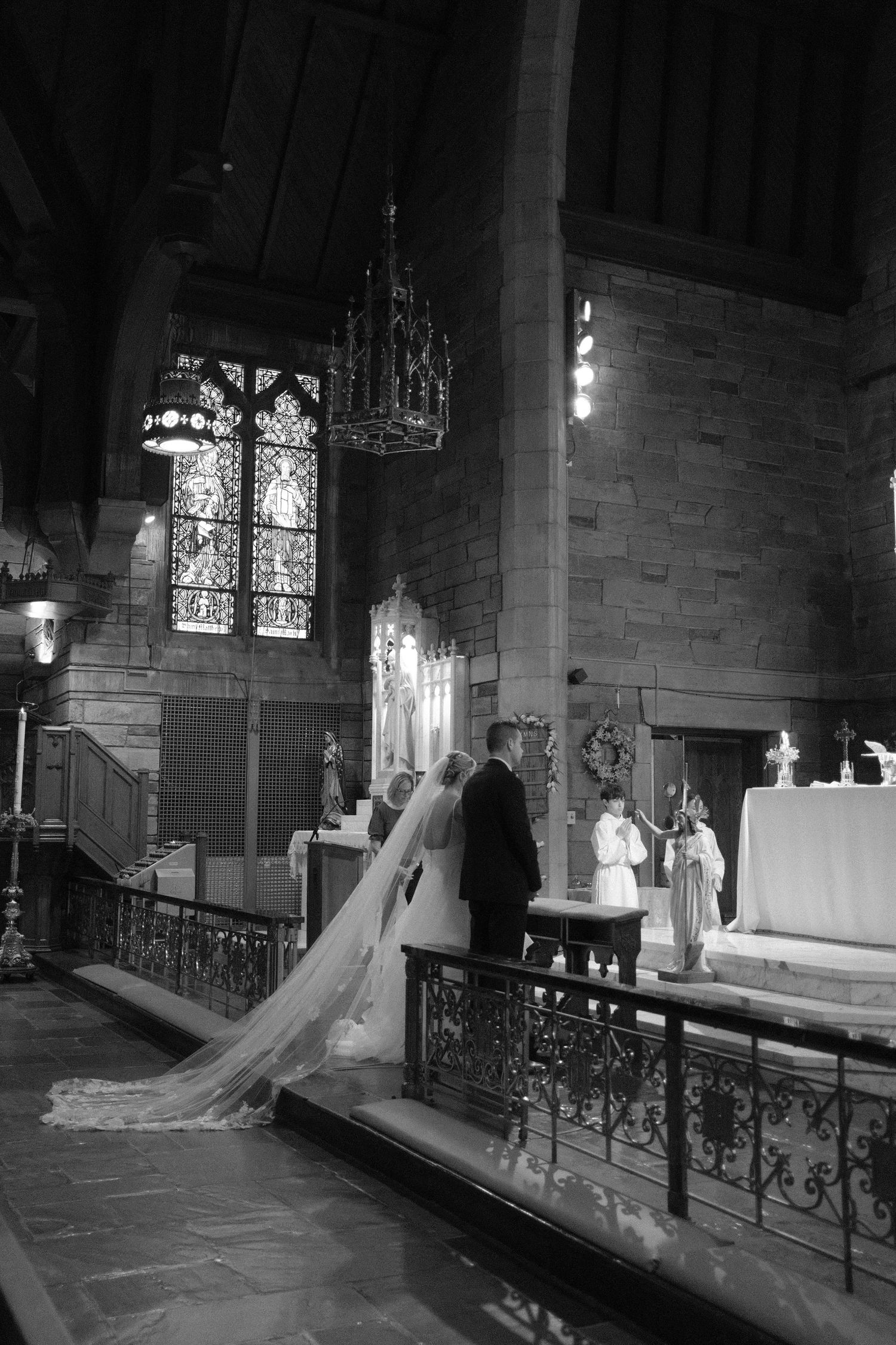 Black and white photo of a bride and groom at a church altar during a wedding ceremony with stained glass windows.