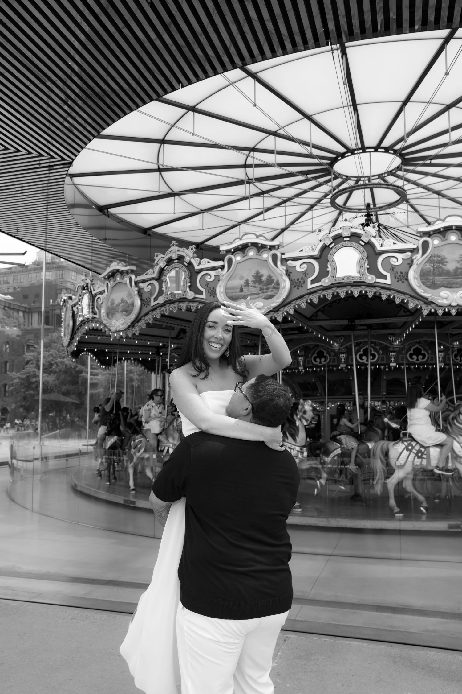 Black and white photo of a couple dancing near a carousel, woman in white dress smiling joyfully.