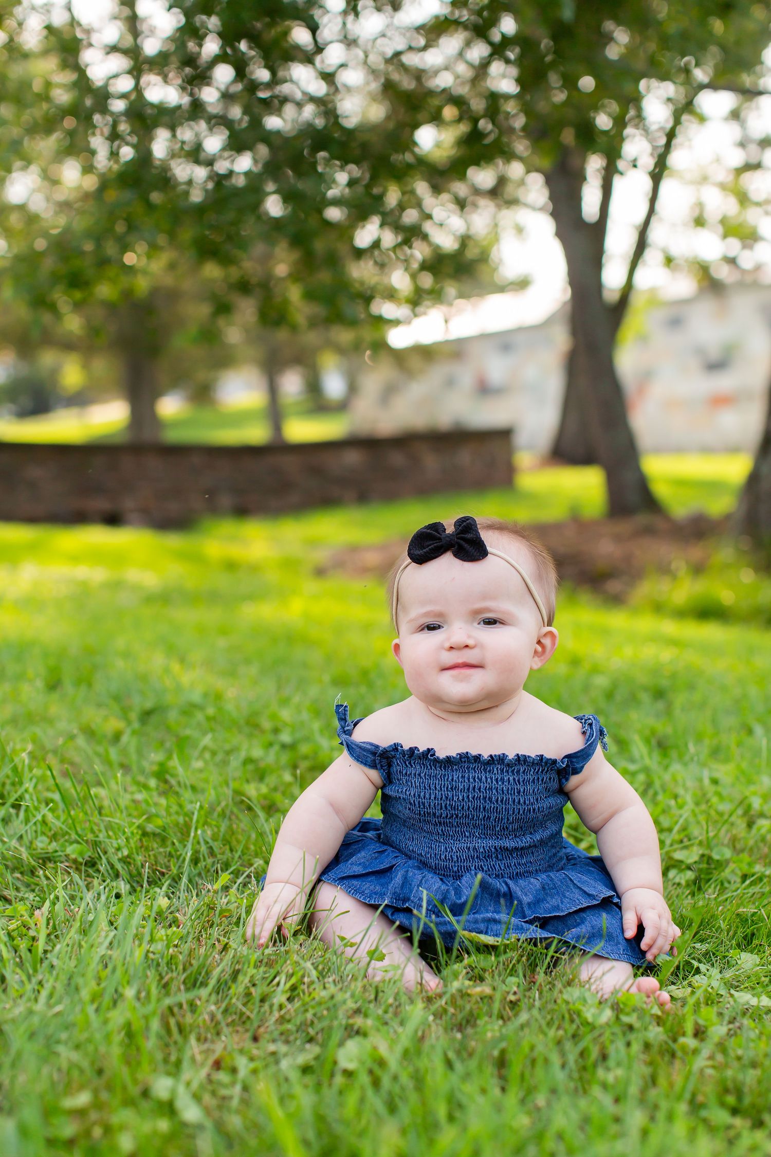 Baby wearing a dark bow and blue outfit sitting in the grass.