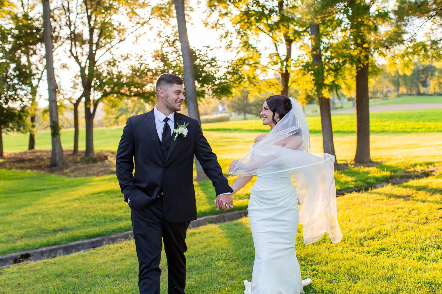 Bride and groom holding hands while walking through sunlit trees.