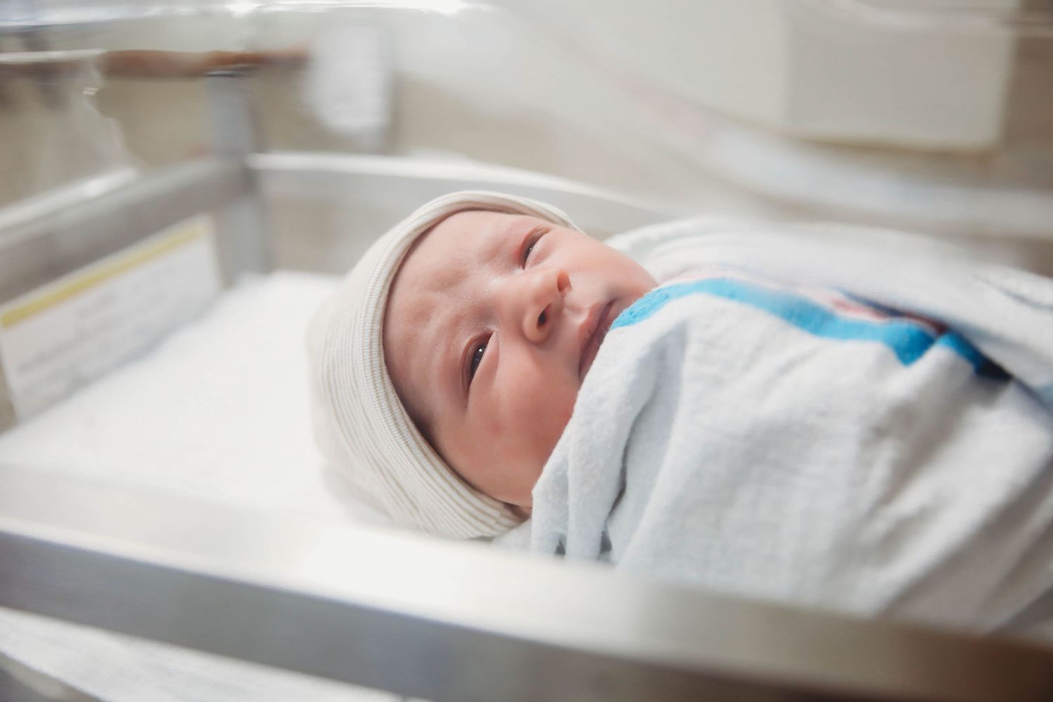 Swaddled newborn wearing a white cap and resting in a hospital bassinet.