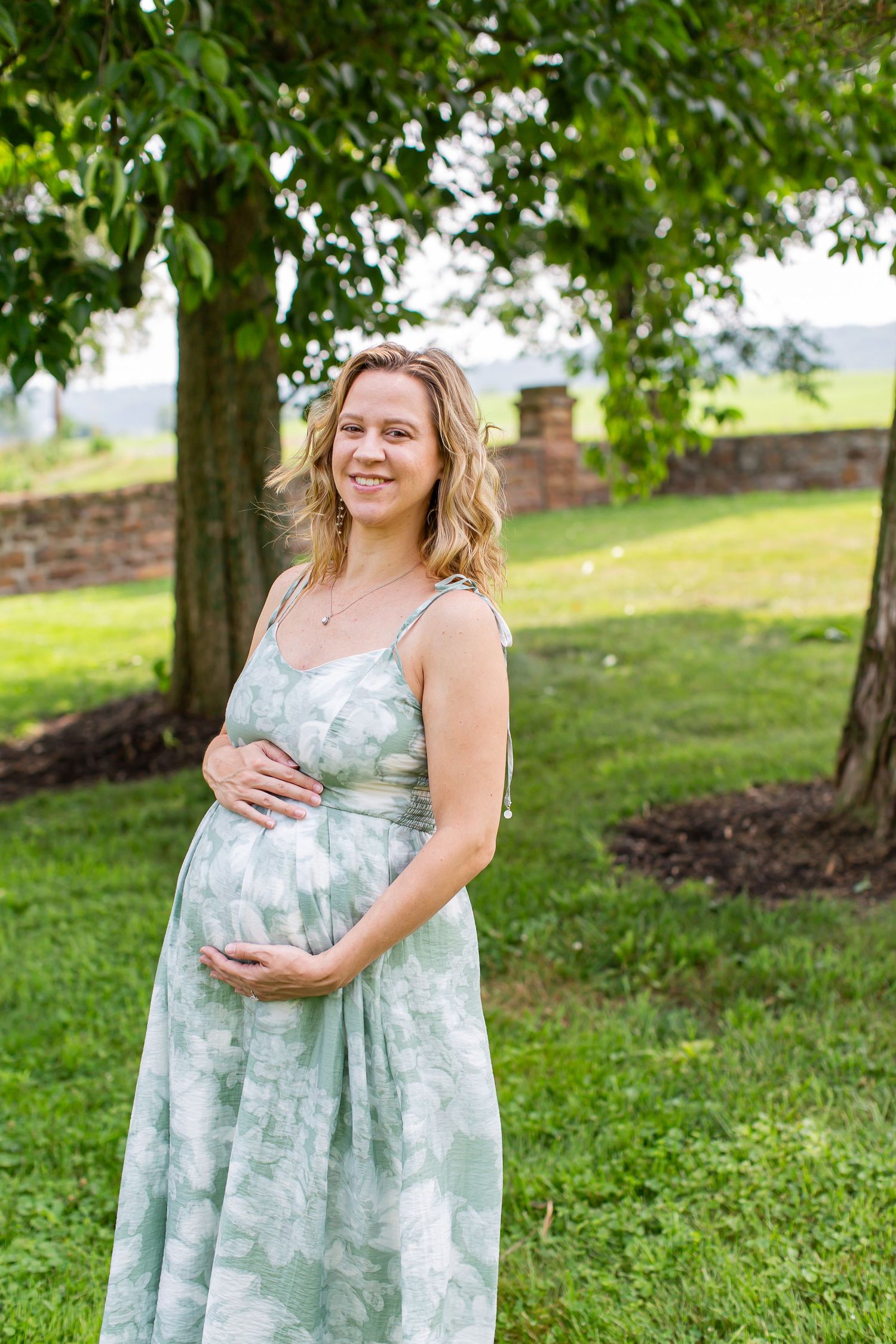 Girl in a pale green dress holding a baby beneath a leafy tree.