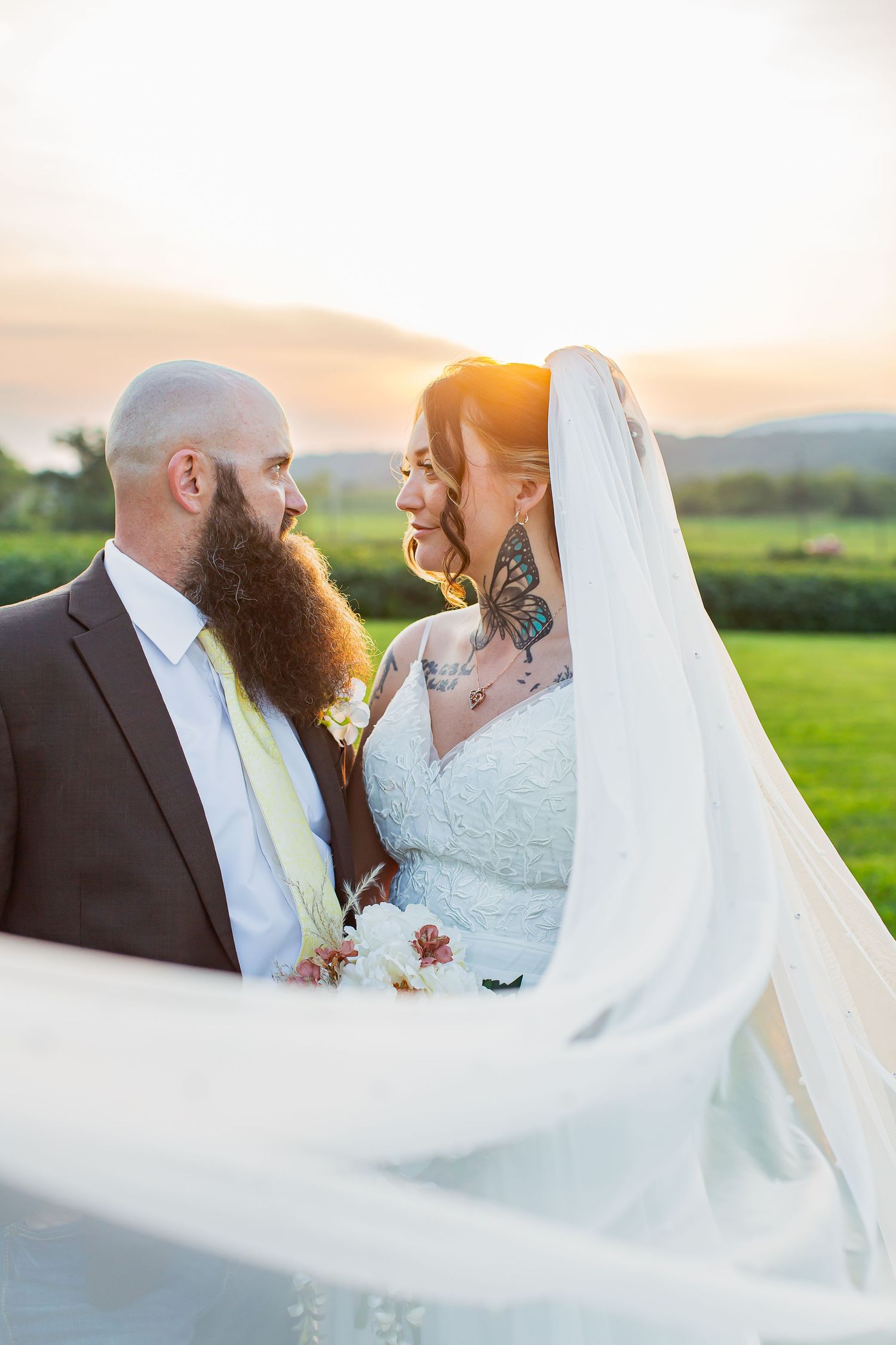 Bride and groom holding hands and smiling at sunset with the bride’s veil in the foreground.