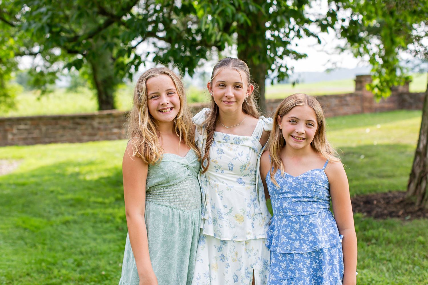 Three girls smiling together outdoors beneath a tree.