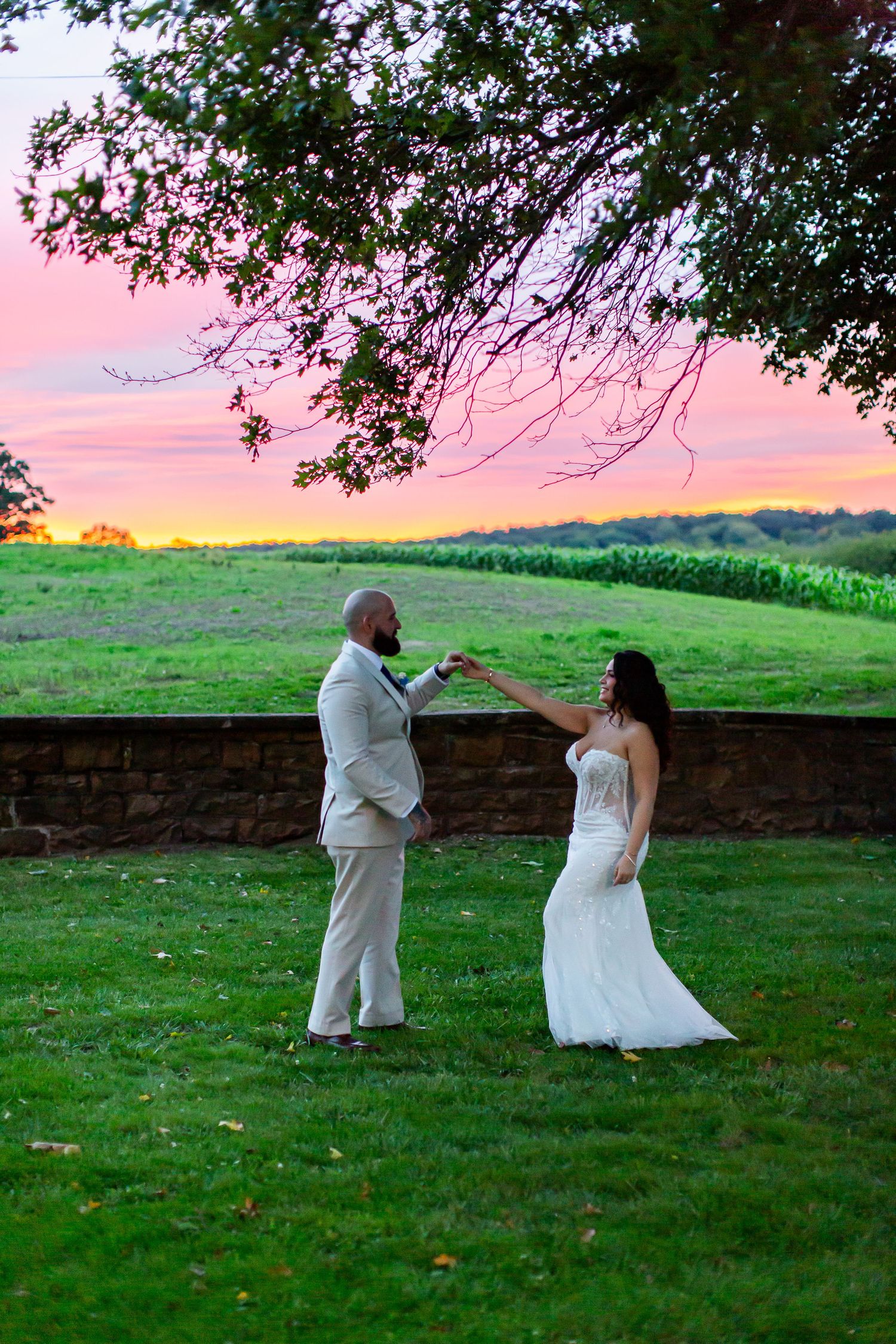 Groom twirling his bride beneath a tree against a pink sunset.
