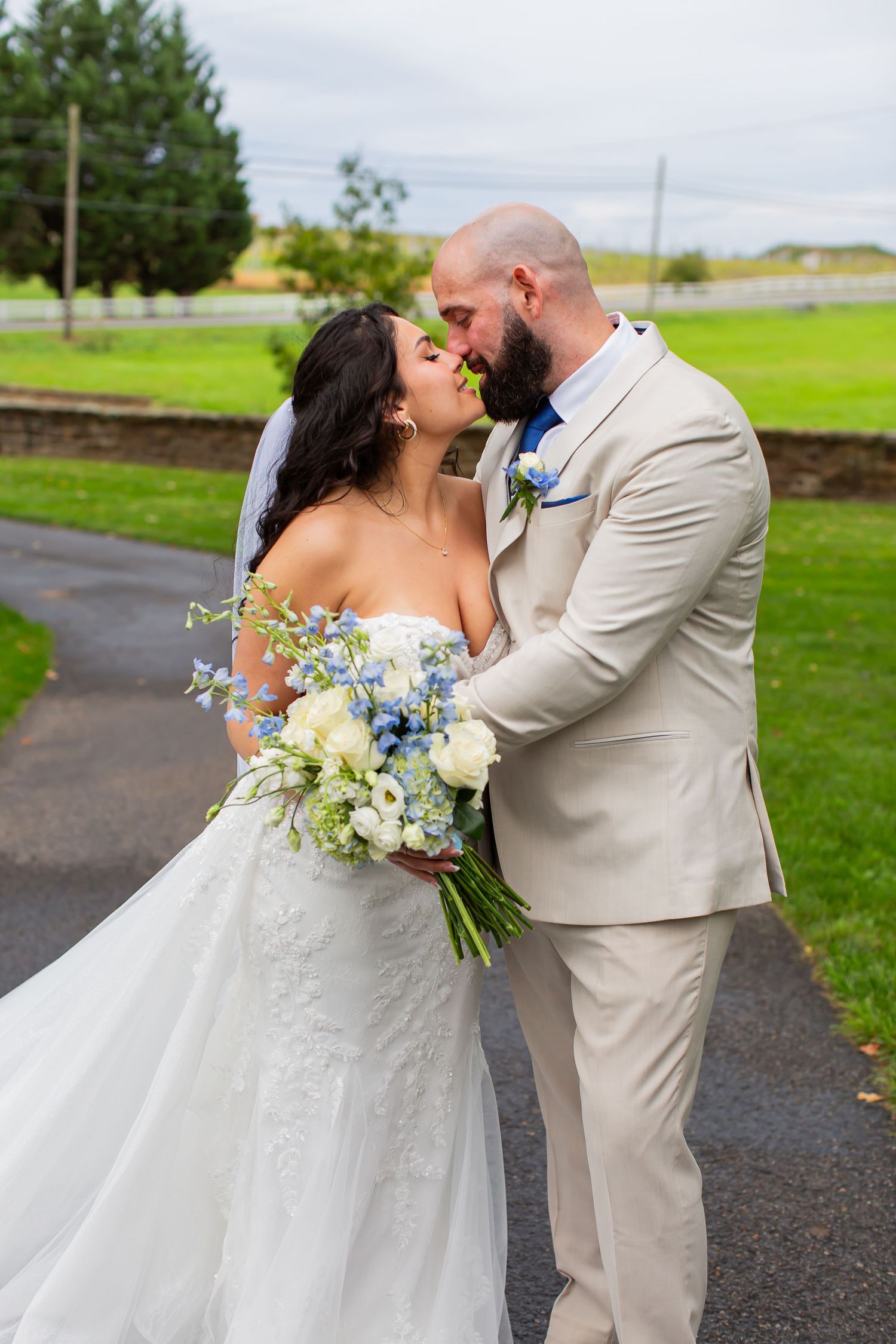 Bride and groom kissing outdoors beside green fields.