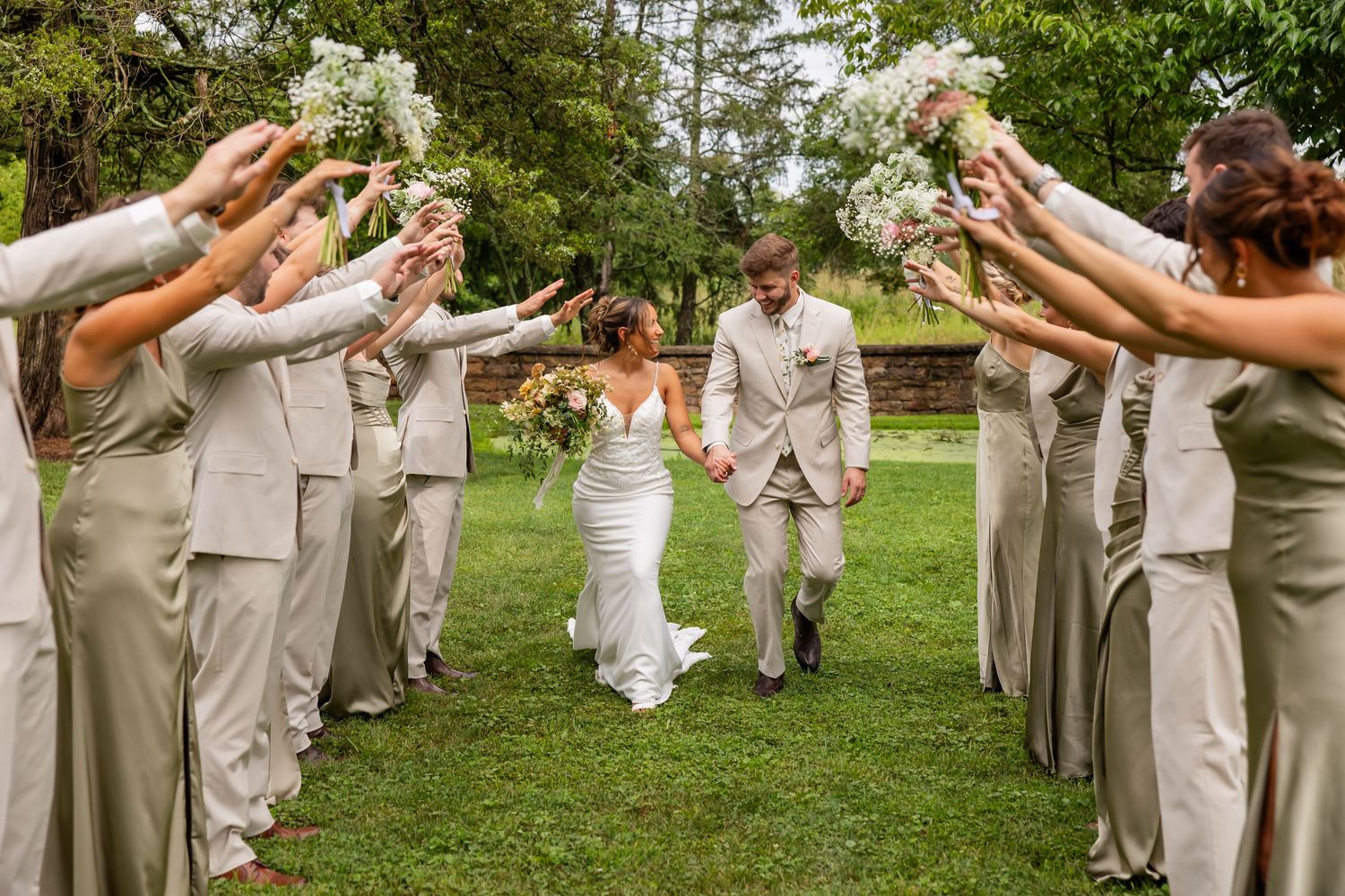 Newlyweds walking between wedding party members raising bouquets outdoors.
