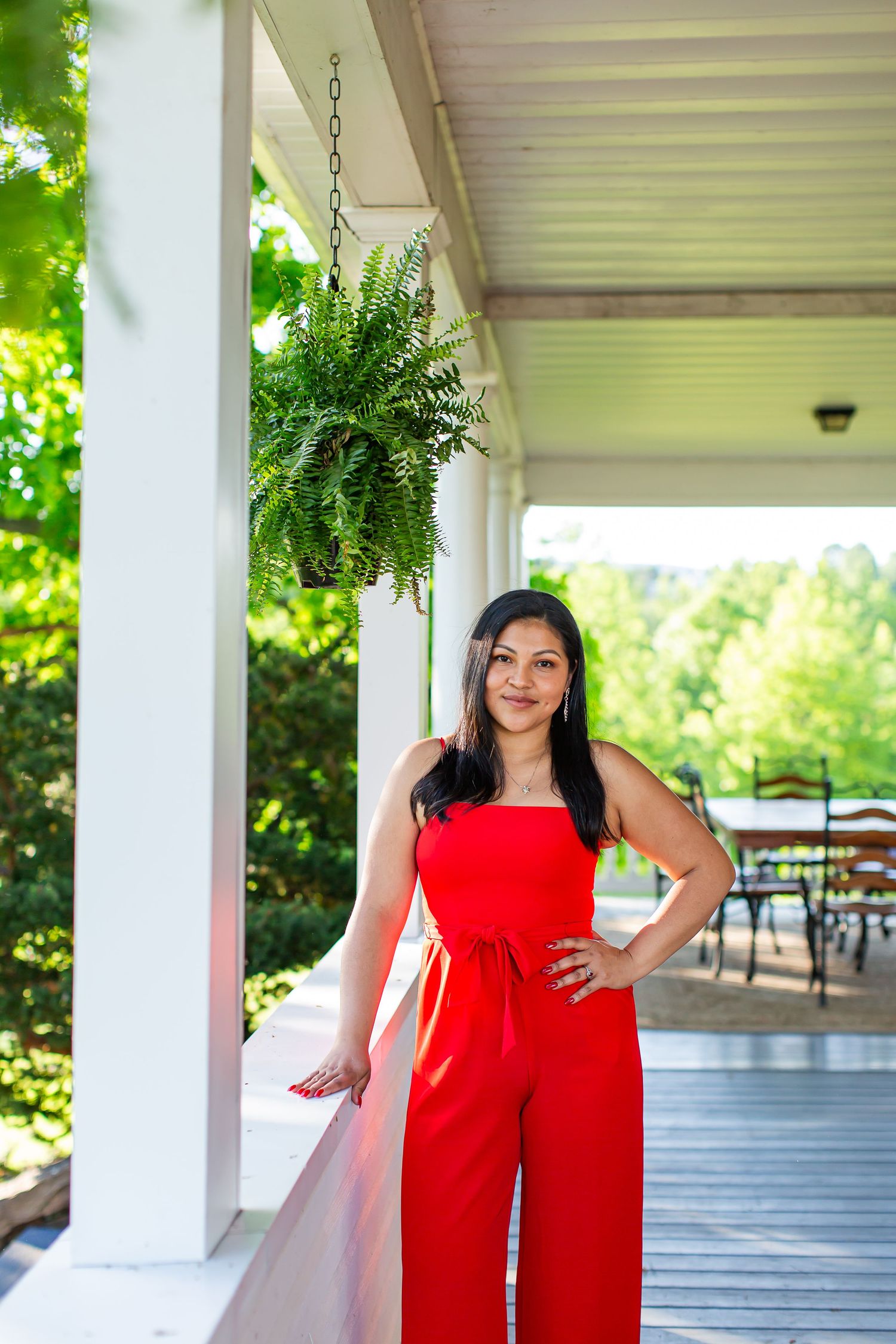 Woman in a red outfit posing on a porch beside hanging greenery.