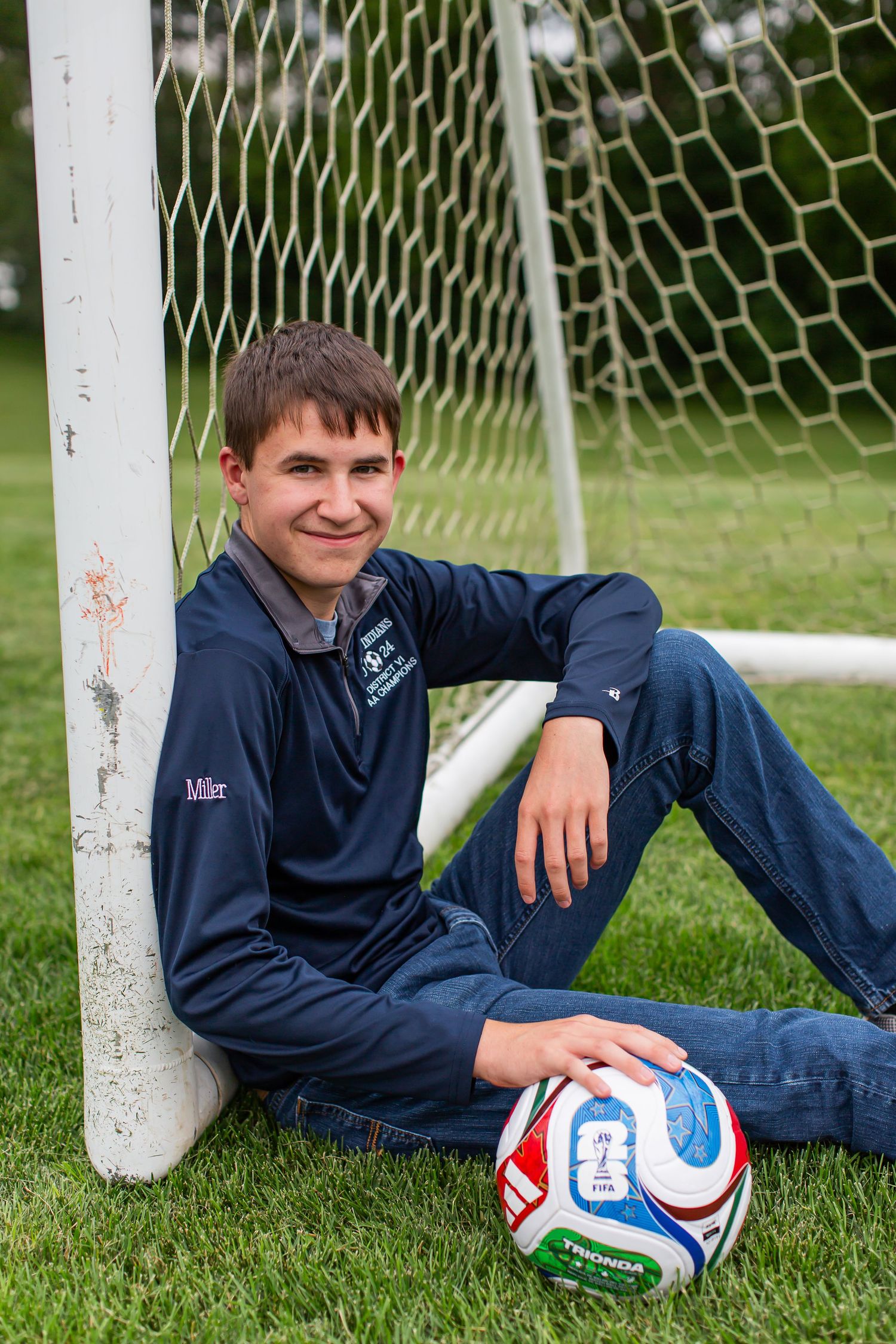 Young man in a navy suit seated beside a soccer goal with a soccer ball.