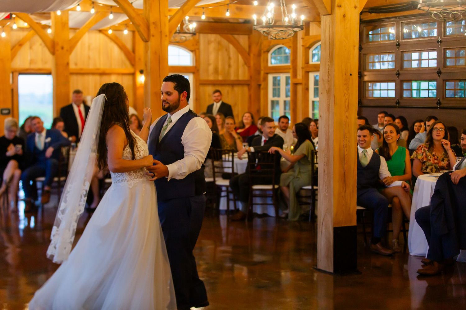 Bride and groom dancing at their wedding reception surrounded by guests and wooden beams.