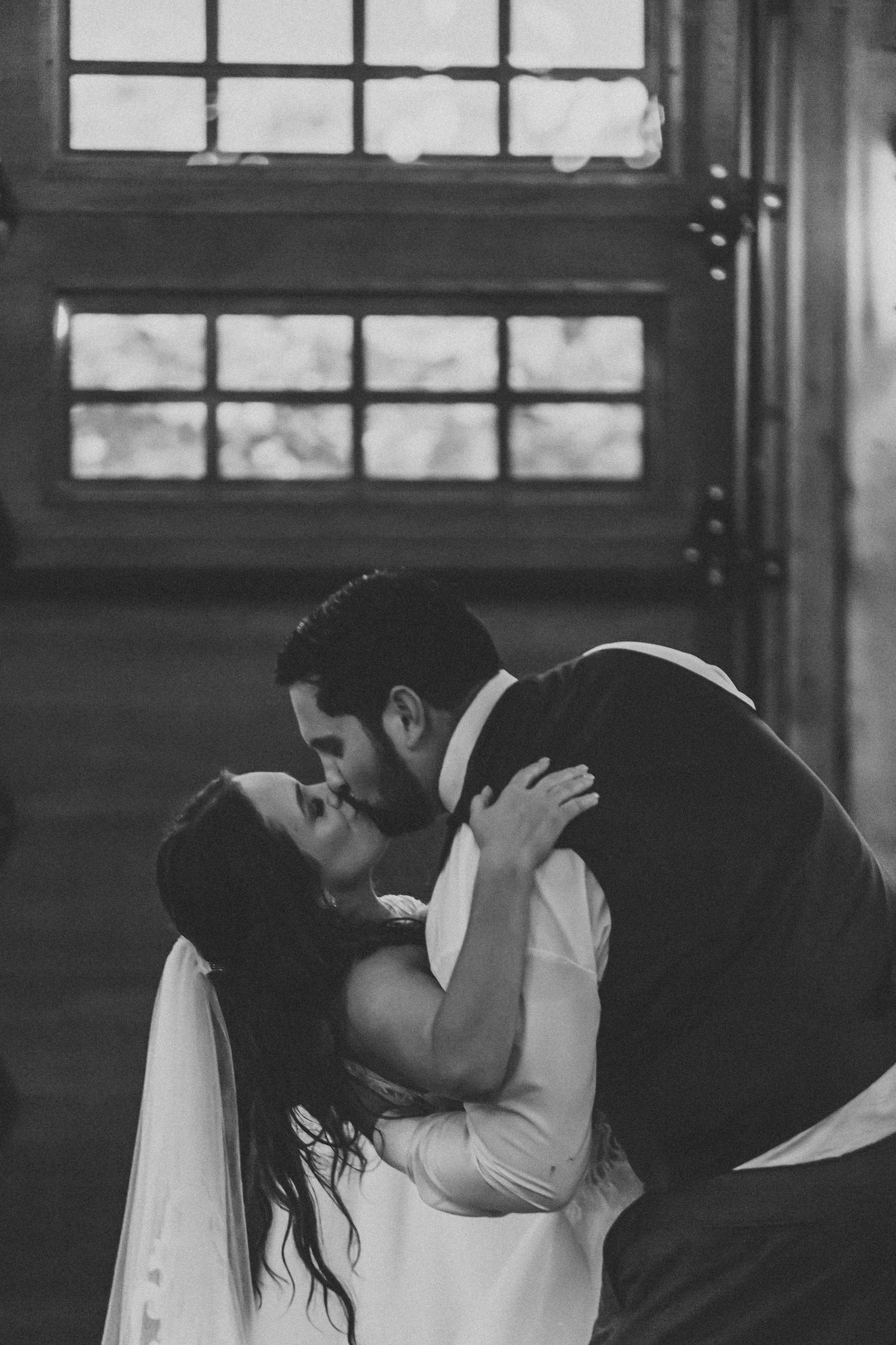 Black-and-white wedding portrait of a groom dipping and kissing his bride beside tall windows.