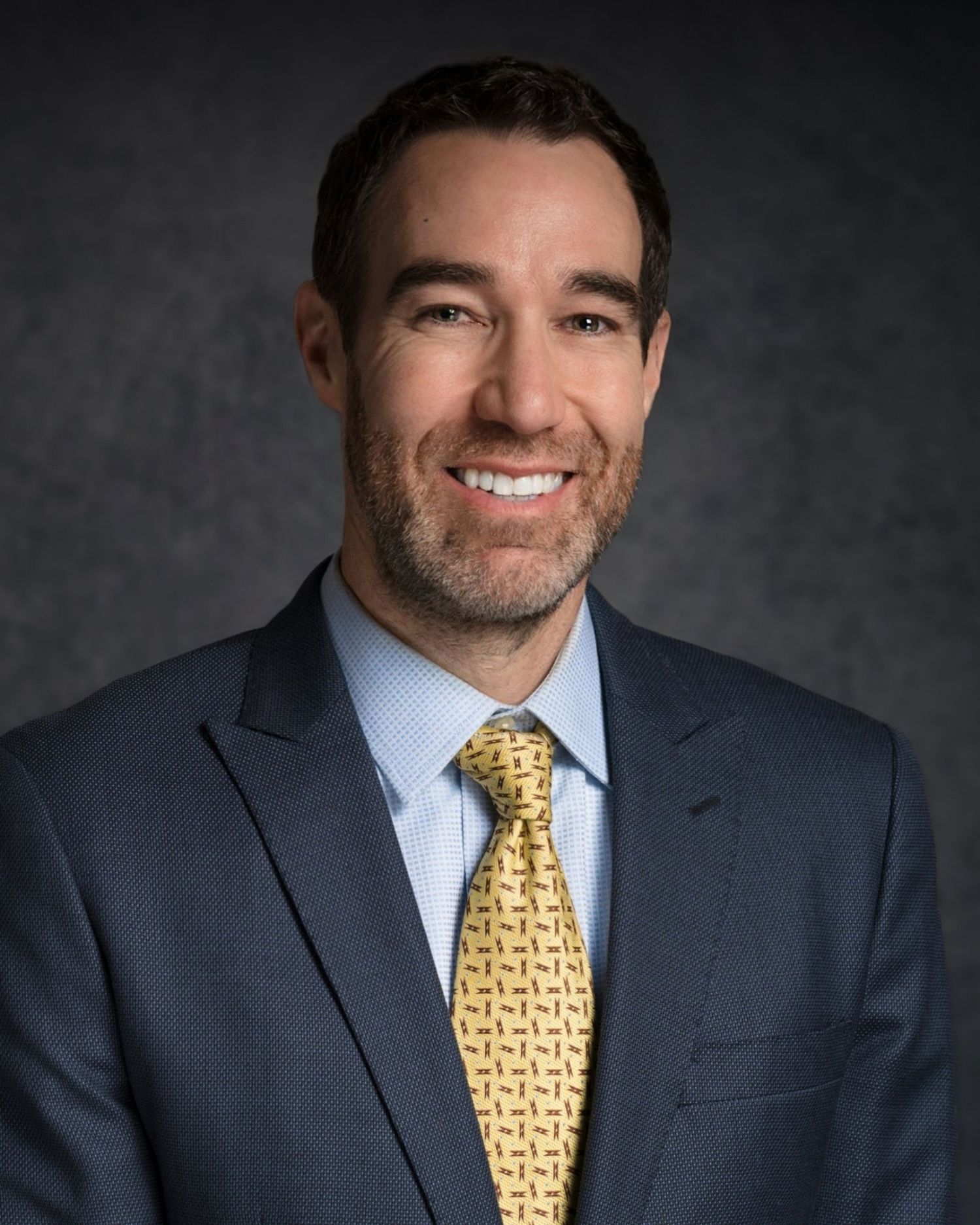 Professional headshot of person in navy suit with yellow patterned tie against dark background.