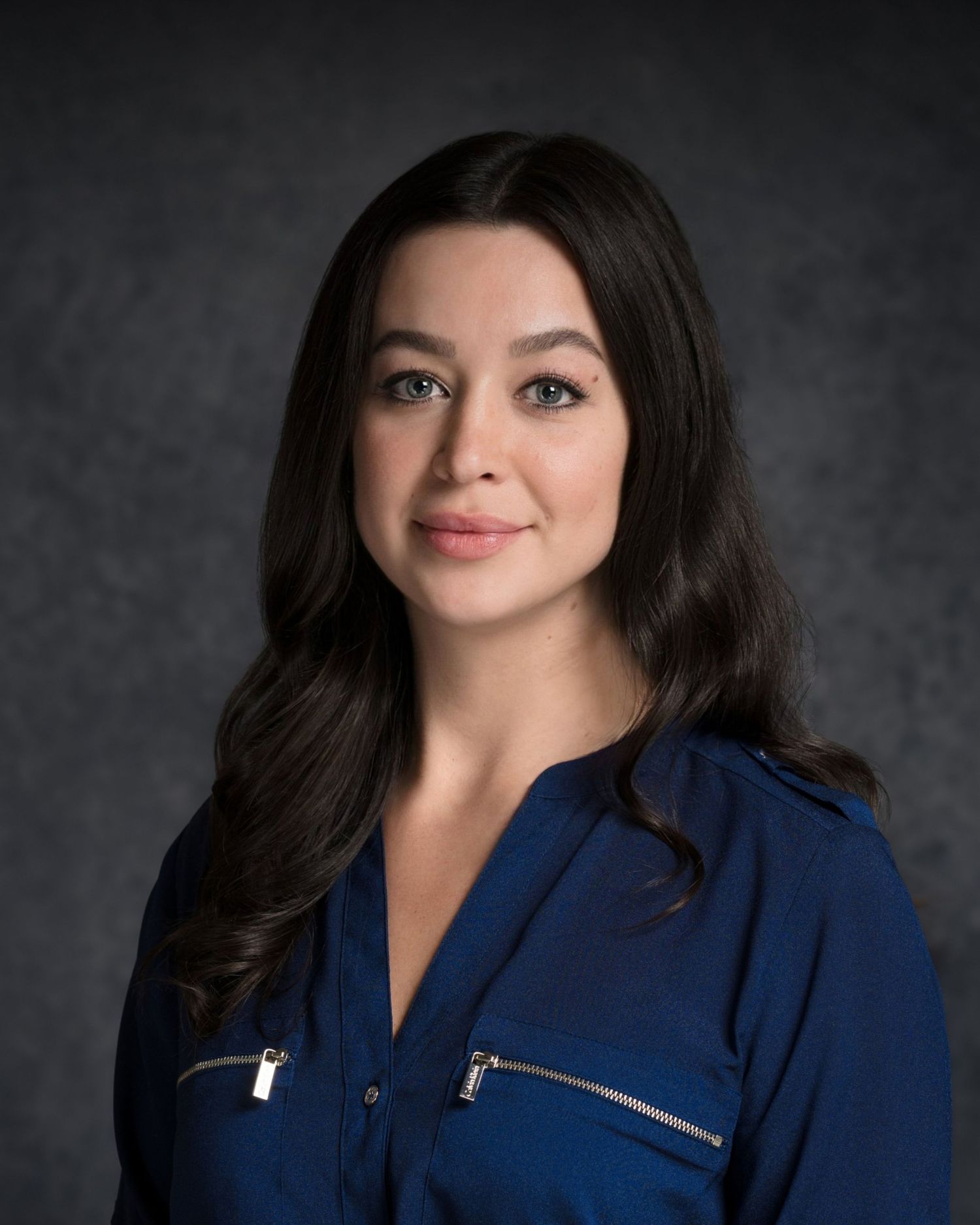 Professional headshot of person in navy blue top with zipper detail against dark background.