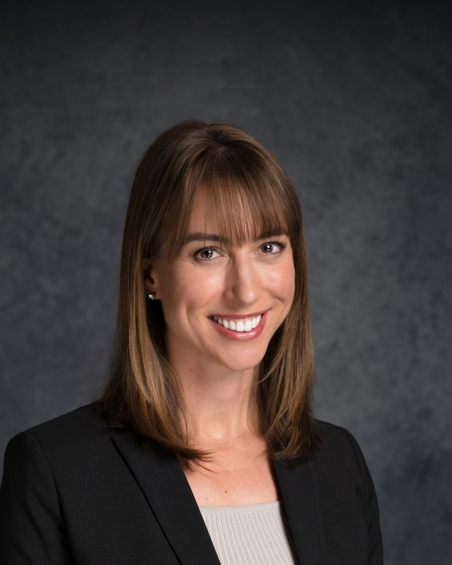 Professional portrait of person in black blazer with grey top against dark backdrop.